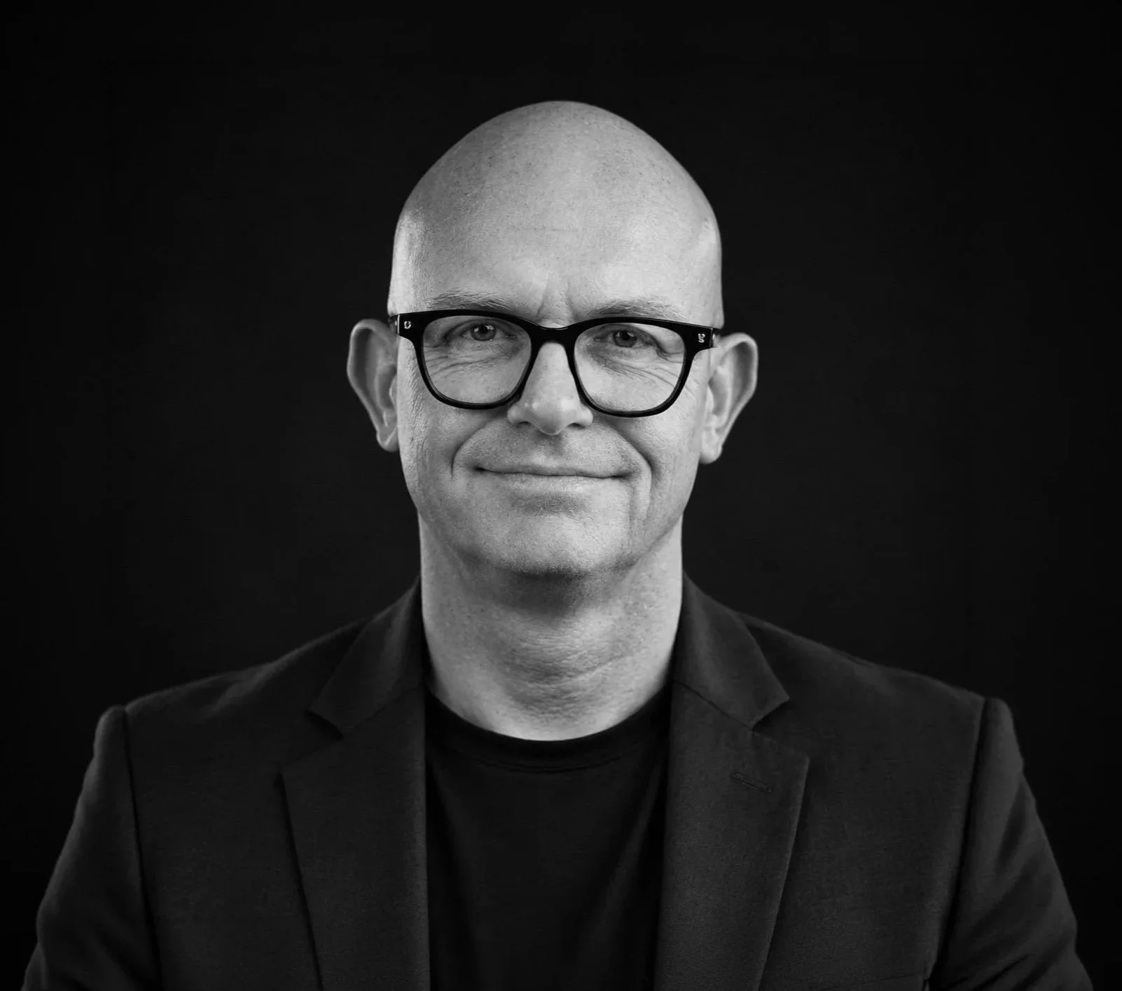 Black and white portrait of a man with a bald head, wearing black glasses and a dark suit, smiling against a black background.