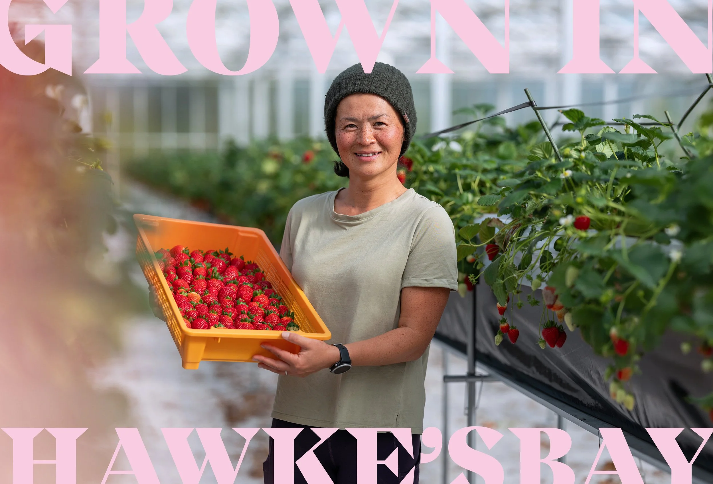 A woman with freckles and a dark beanie smiles while holding a large orange basket of fresh strawberries inside a greenhouse.