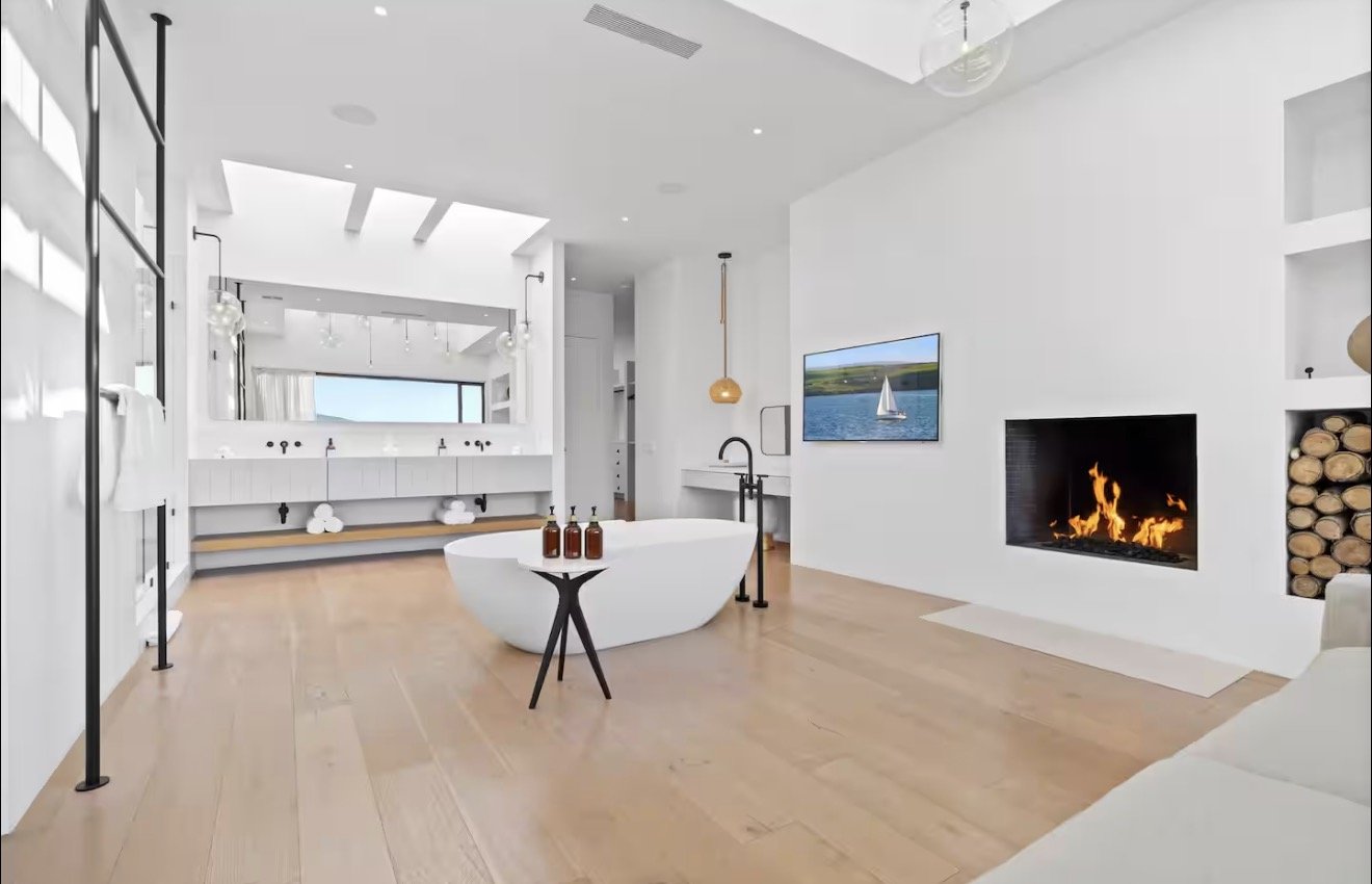 Modern living room with white walls, a fireplace with flames, a wall-mounted TV showing a sailboat on water, a freestanding bathtub, a small black side table with bottles, a large mirror above double sinks, hardwood floors, and minimalistic decor.