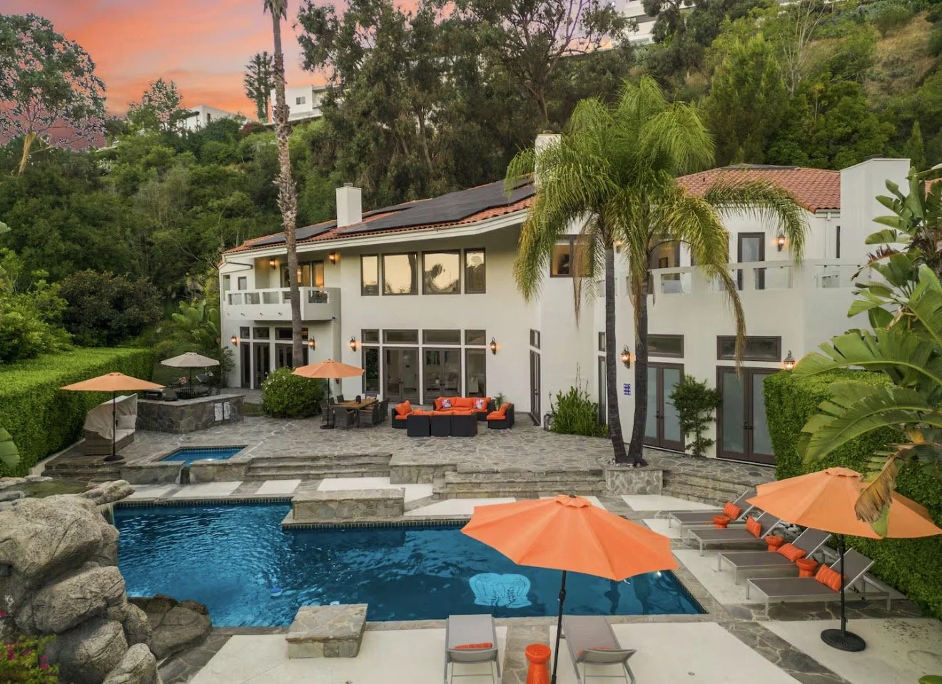 Luxury house with large patio and swimming pool, surrounded by greenery and trees, during sunset.