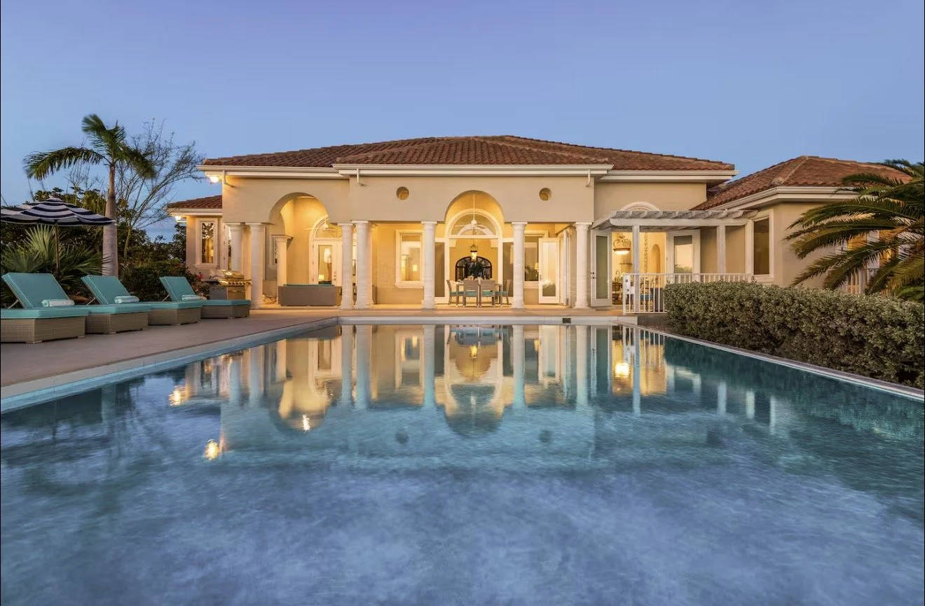 Luxury house with a swimming pool in the front yard during dusk, featuring outdoor lounge chairs and palm trees.