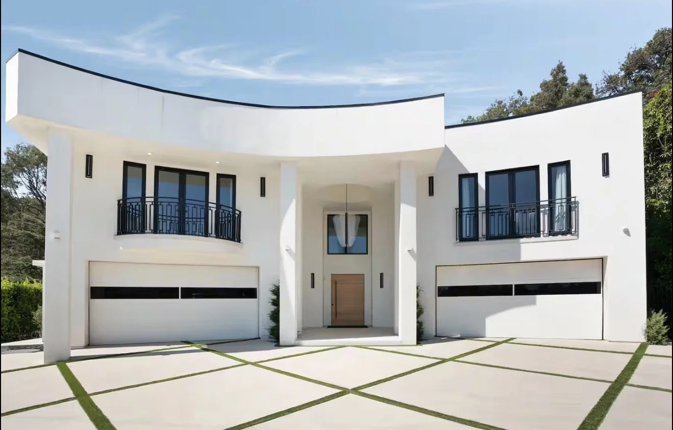 Modern white two-story house with black window frames and balconies, garage doors, and a unique curved roof design, surrounded by greenery.