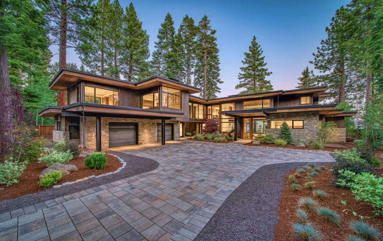 Modern house with large windows, stone and wood exterior, surrounded by trees, with a paved driveway and landscaped garden.