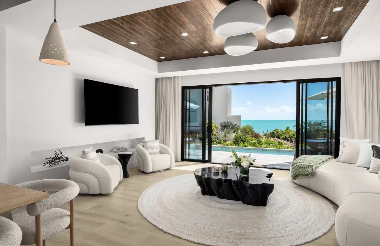 Modern living room with white furniture, a black coffee table, sliding glass doors opening to a view of the ocean, and a wooden ceiling.
