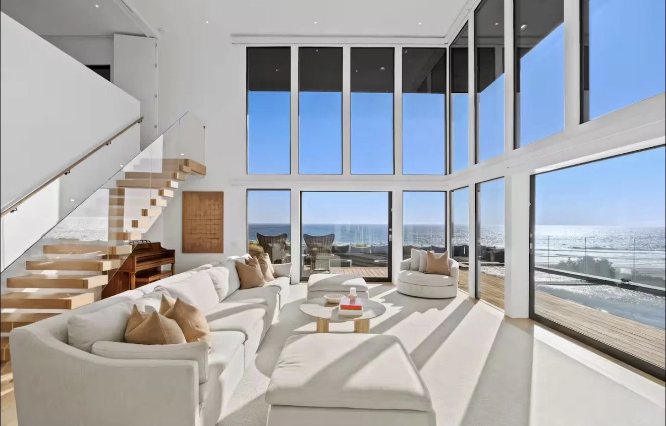 Modern living room with large floor-to-ceiling windows overlooking an ocean view, white furniture, a staircase, and bright sunlight.