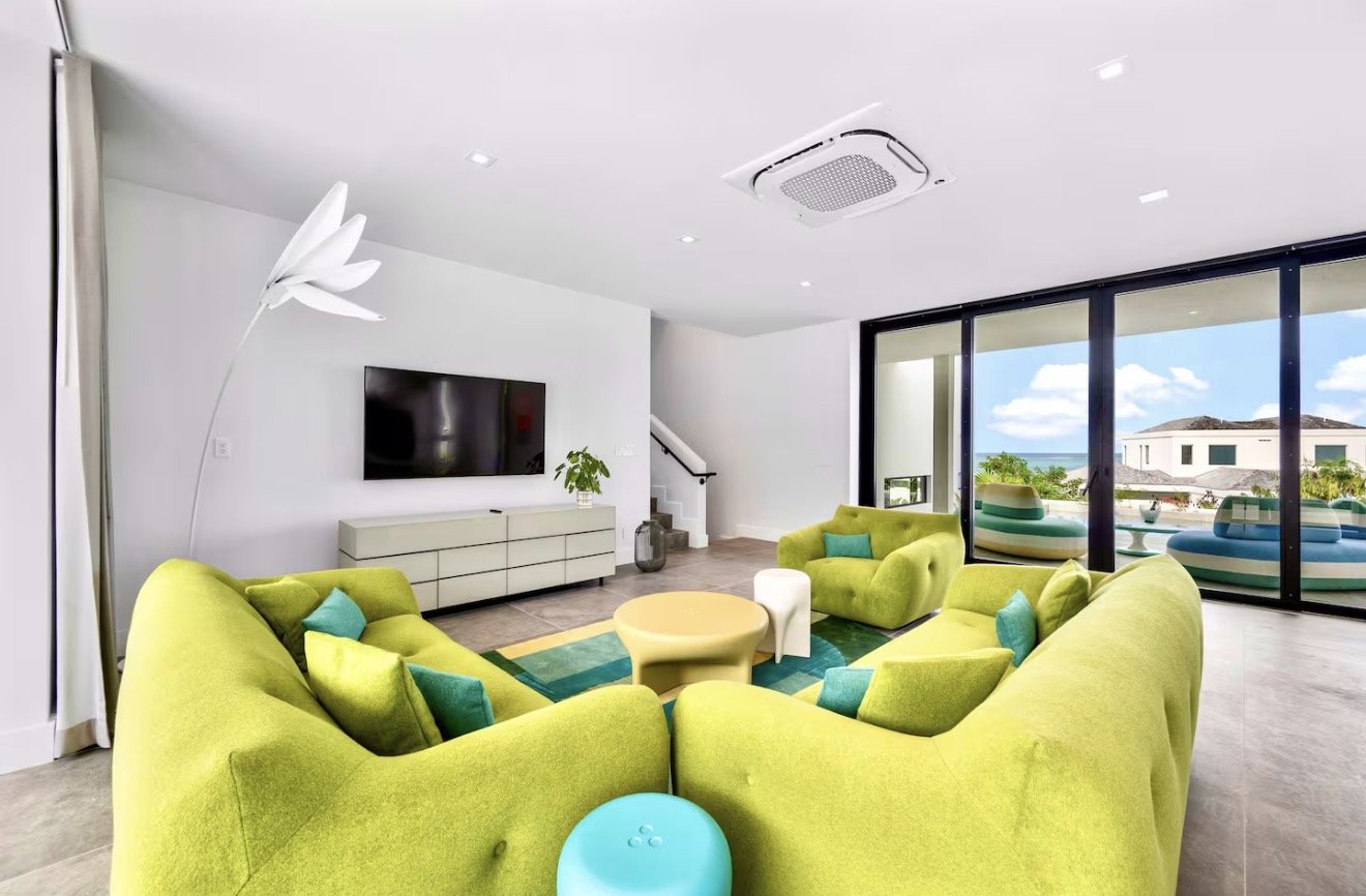 Living room with lime green couches, a flat-screen TV, a white sideboard, a modern white floor lamp shaped like a flower, large windows showing a view of the ocean, and a small balcony with colorful outdoor chairs.