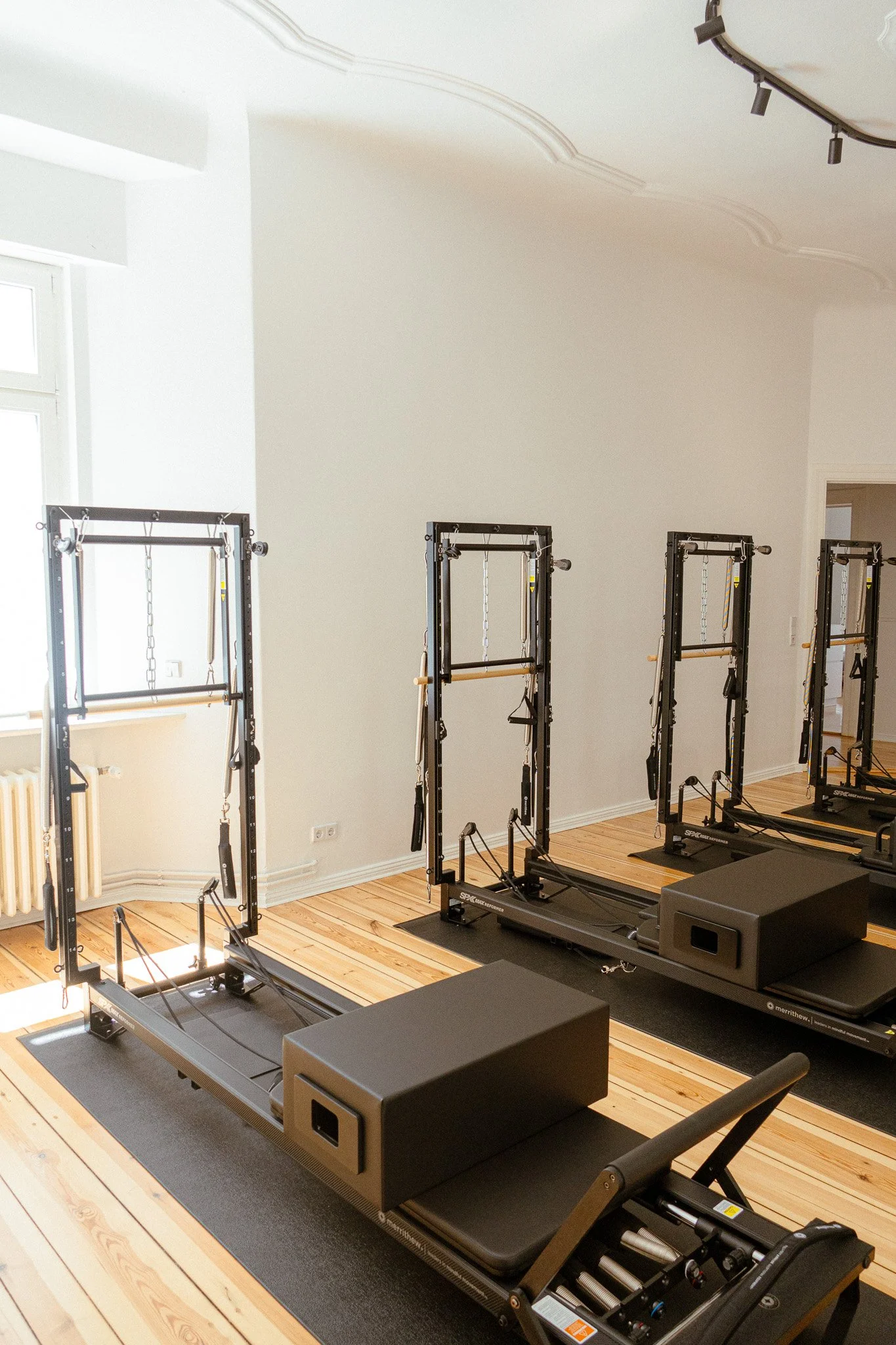Der Geräte-Raum mit 4 Reformern, Holzboden und weißen Wänden, natürliche Beleuchtung durch Fenster. An der Decke sieht man etwas Stuck von dem Altbau und einen Teil der Lampe.