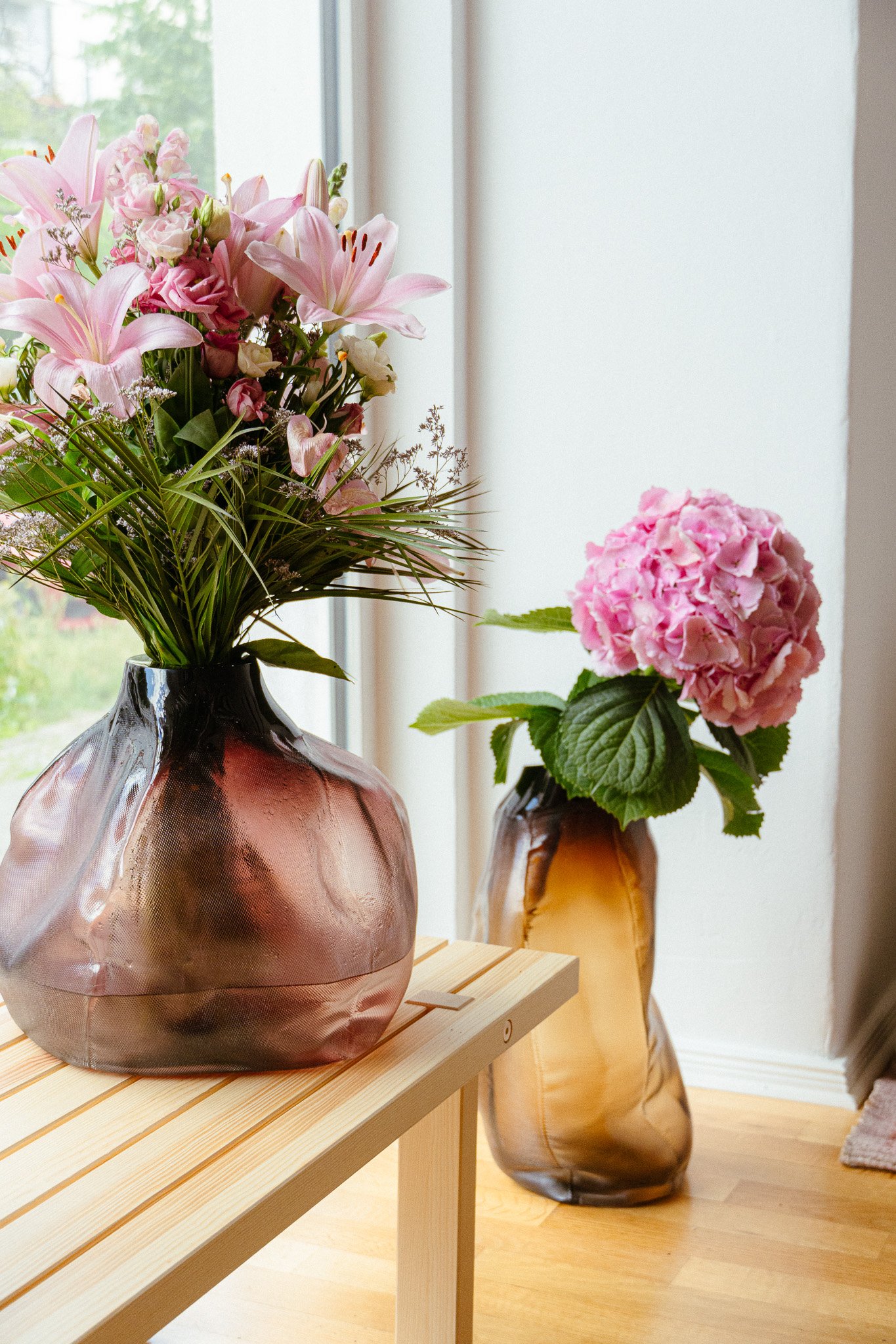 Zwei Blumenvasen mit Blumen, eine auf einem Holztisch, die andere im Hintergrund, in einem hellen Raum mit Holzfußboden.