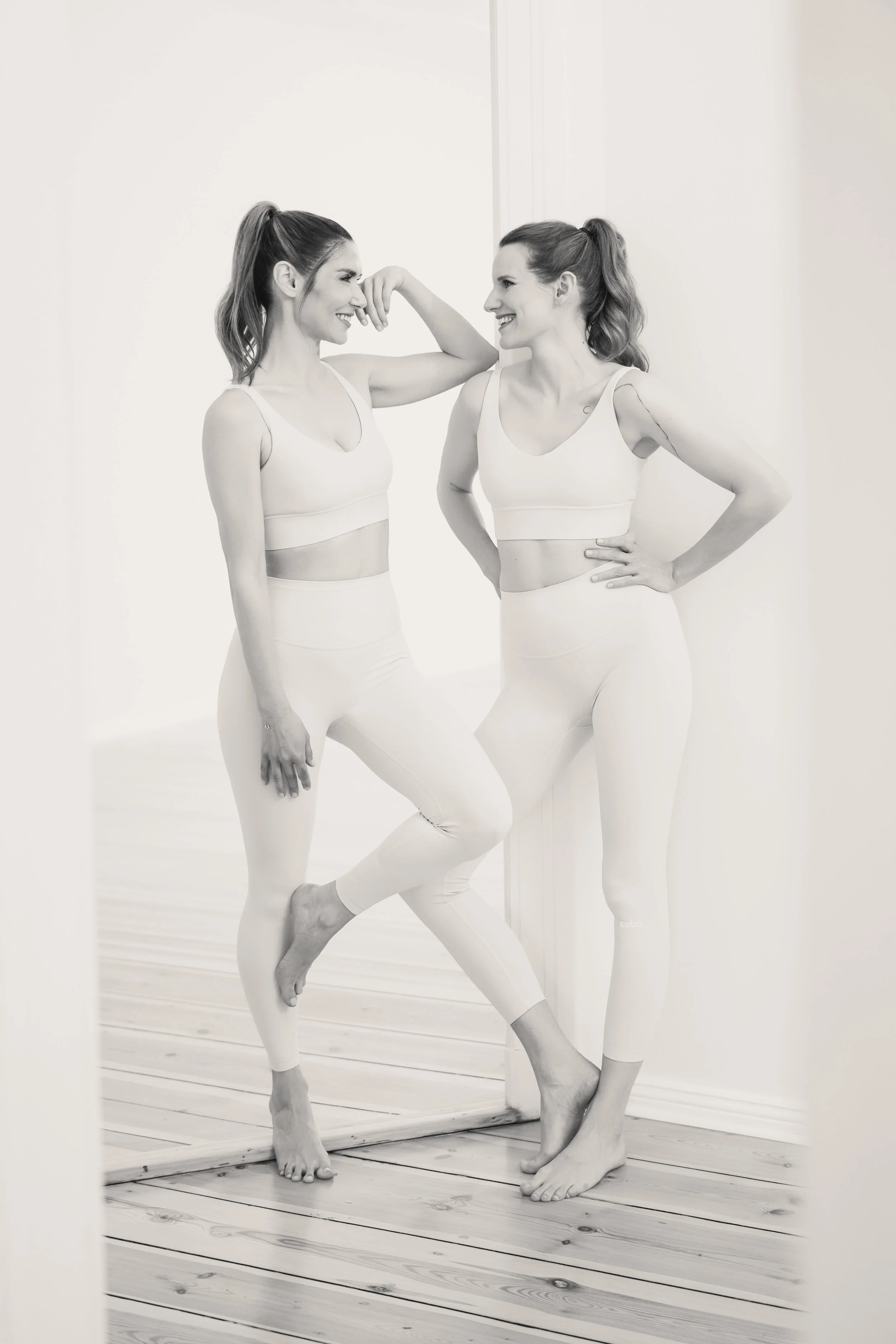 Mimi und Lucie in Sportkleidung, schauen sich lachend an, stehen auf Holzparkettboden vor weißer Wand.