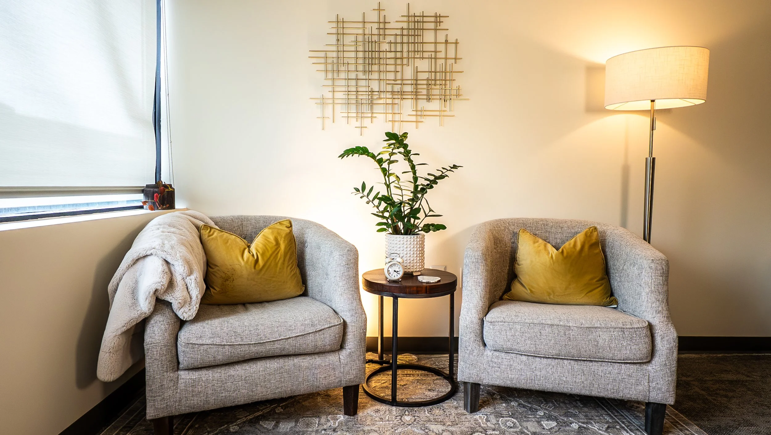 Living room with two beige armchairs featuring yellow pillows, a small round wooden table with a potted plant and a clock, a floor lamp, a window with a blind, and decorative wall art.