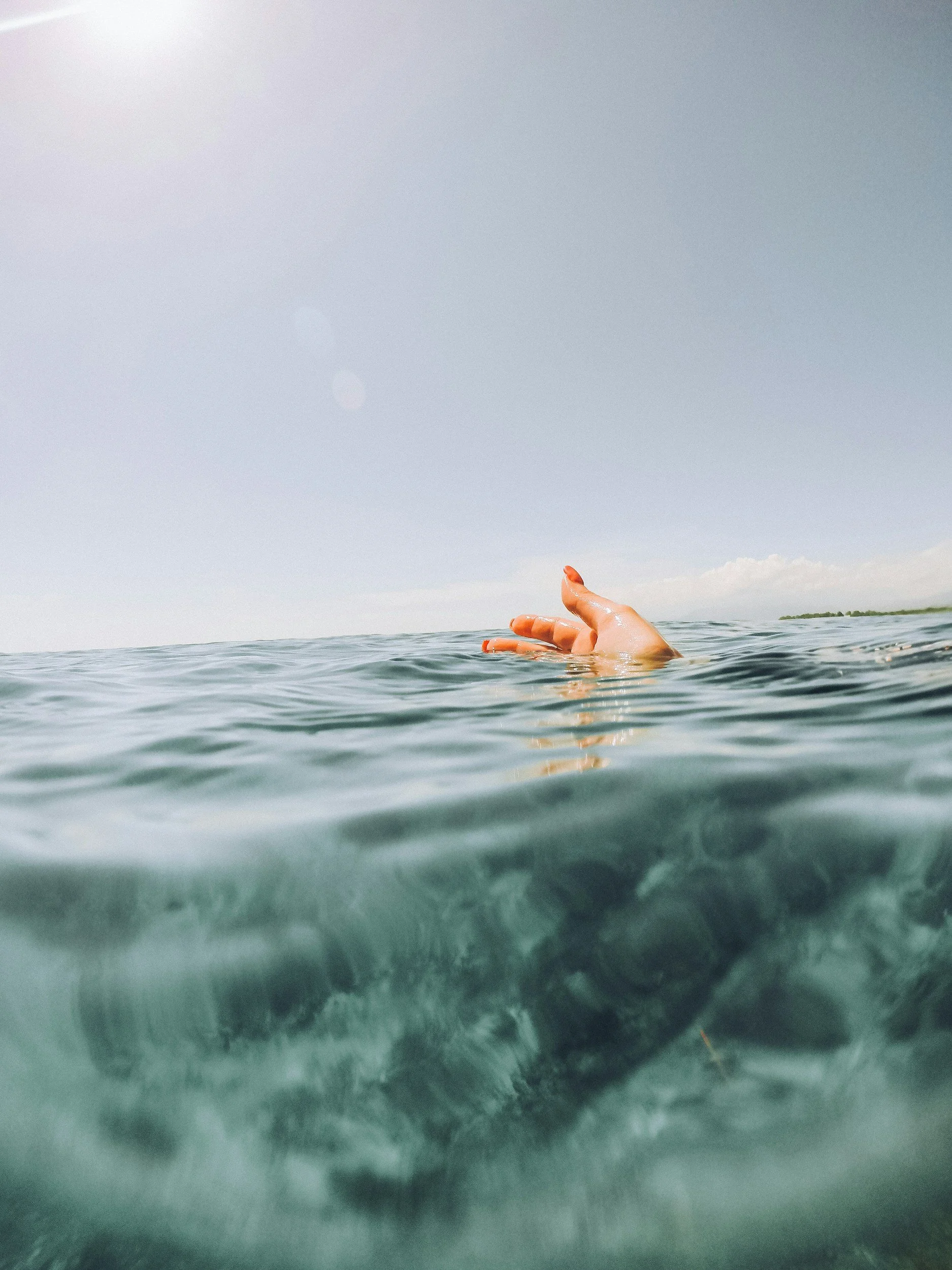 A person swimming in open water, with part of their hand visible above the surface and a distant shoreline against a cloudy sky.