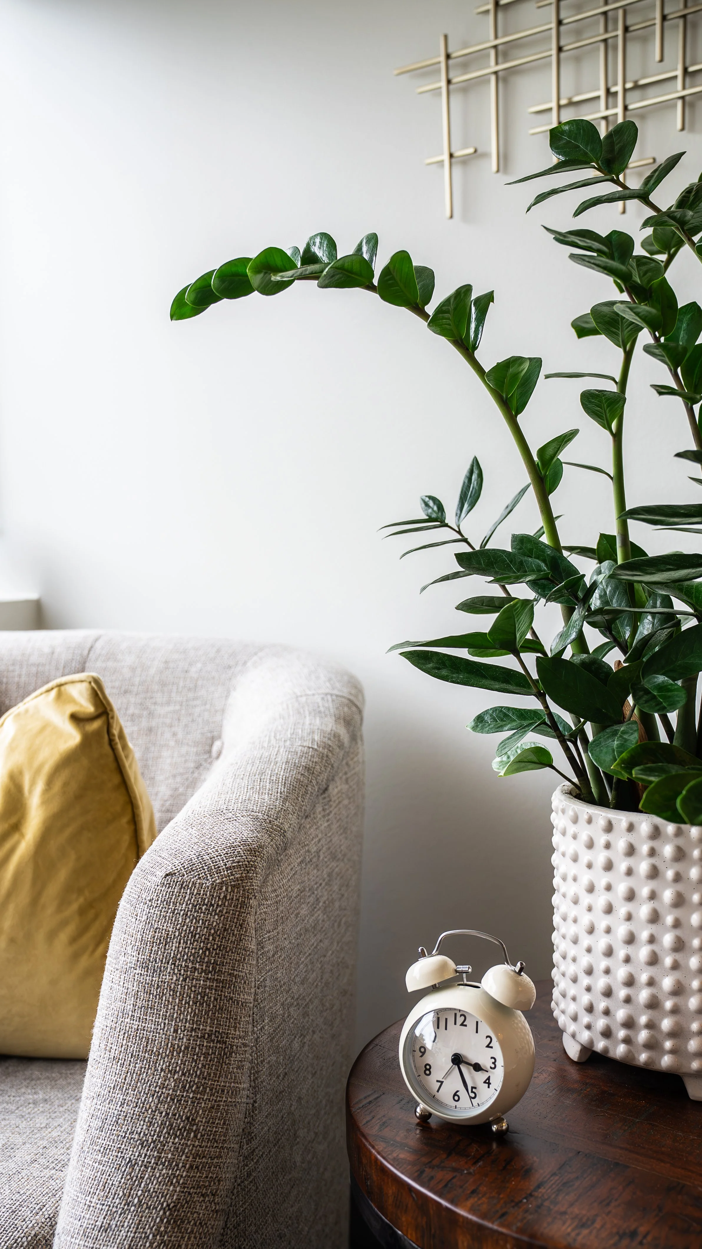 A living room scene with a beige upholstered sofa, a mustard yellow pillow, a round wooden side table, a white ceramic pot with a large green houseplant, and a white vintage alarm clock showing 4:09. A minimalist wall art piece is on the white wall in the background.