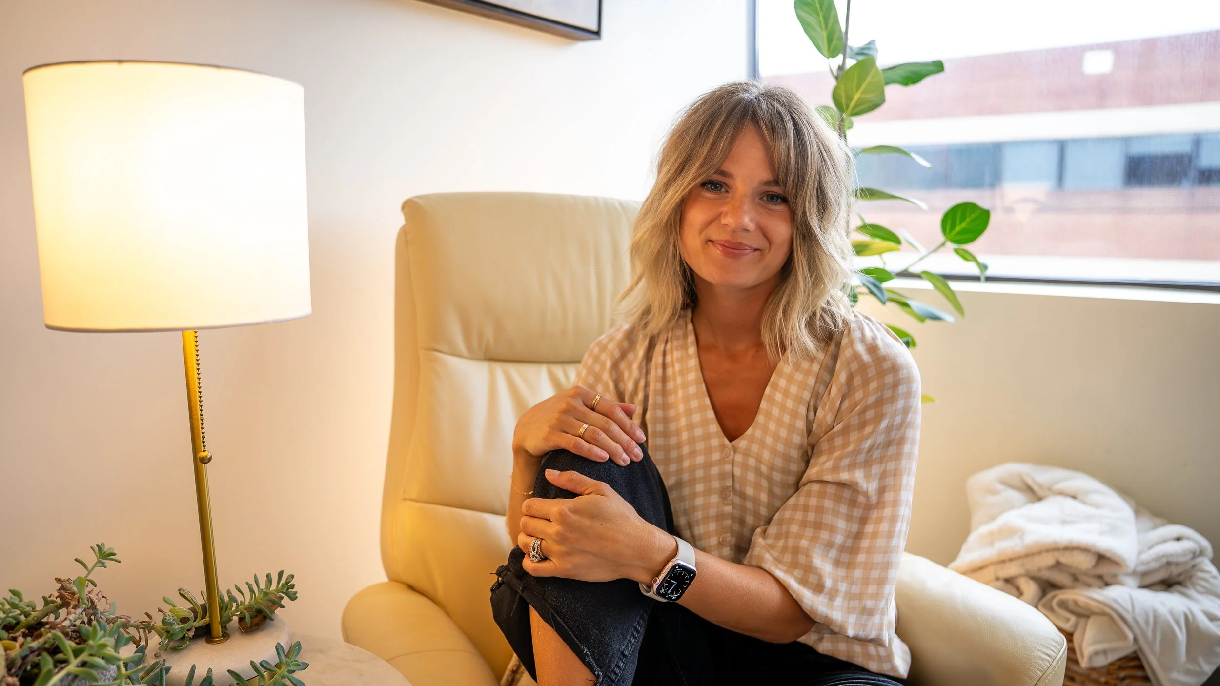 A woman with blonde wavy hair sitting on a cream-colored chair near a window, smiling softly, wearing a beige checkered shirt, with a potted plant and a table lamp next to her.