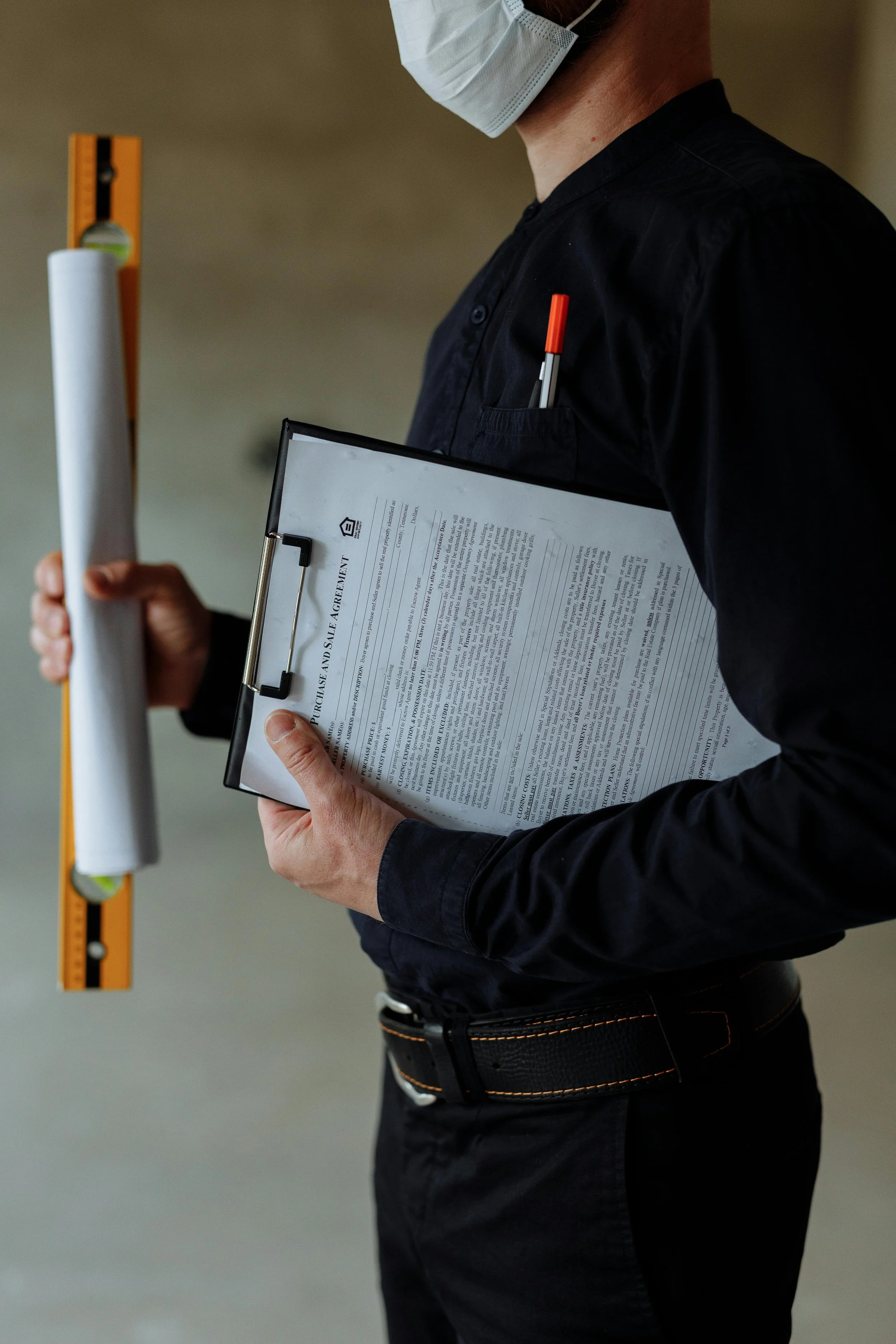 Persoon met zwart shirt en zwarte broek houdt een clipboard met documenten en een pen, draagt een wit gezichtsmasker en heeft een pen in de borstzak. Naast persoon staat een meetlint en een waterpas.