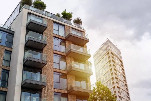 Twee moderne hoog gebouwen met balkons en groene planten op het dak, onder een bewolkte hemel.