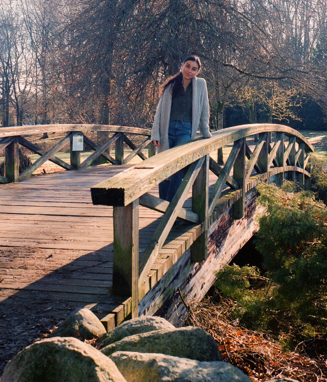 A woman wearing a gray cardigan and dark clothing stands on a small wooden bridge in a park during late fall or winter, with leafless trees in the background and sunlight casting long shadows.
