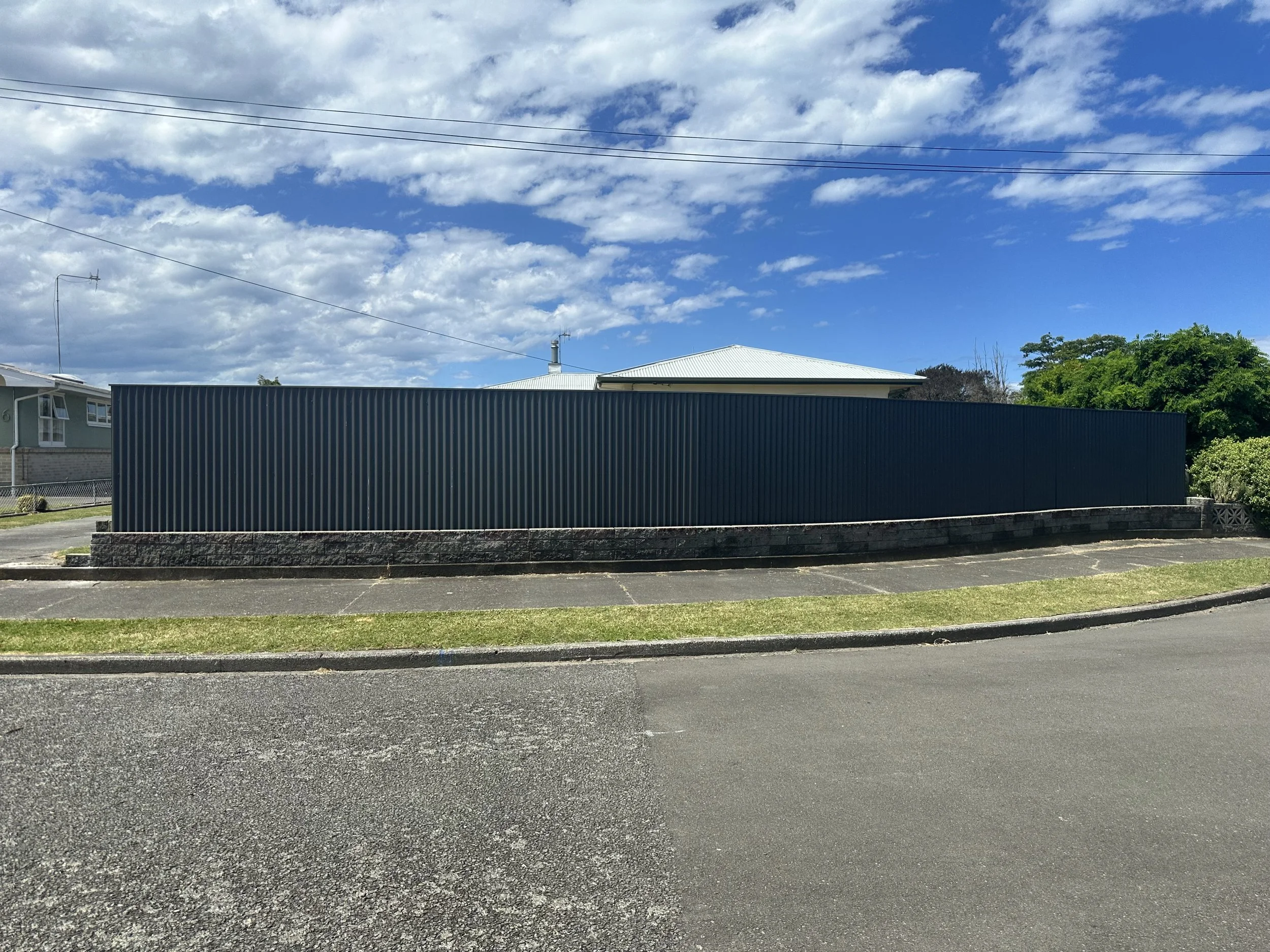 A residential street with a tall, dark-colored colour steel fence, a sidewalk, and grass with a curb in front. There are houses in the background and a sky with scattered clouds.
