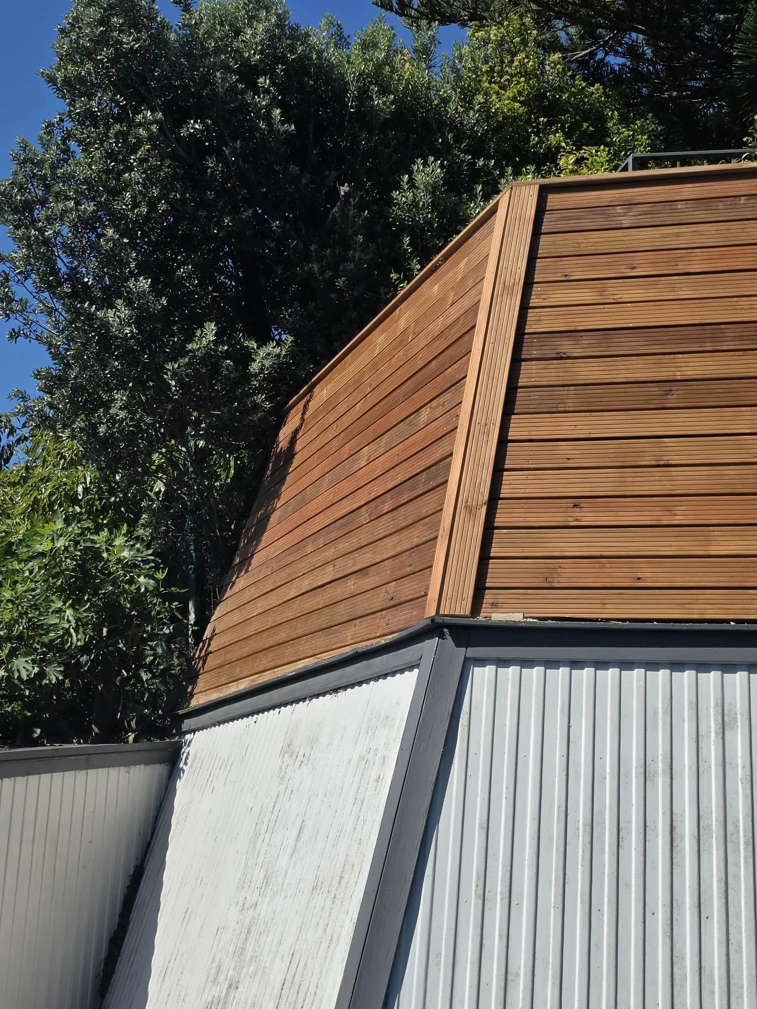 Close-up view of a building's exterior featuring a roof with wooden panels and a section with corrugated metal siding, with green trees and a clear blue sky in the background.