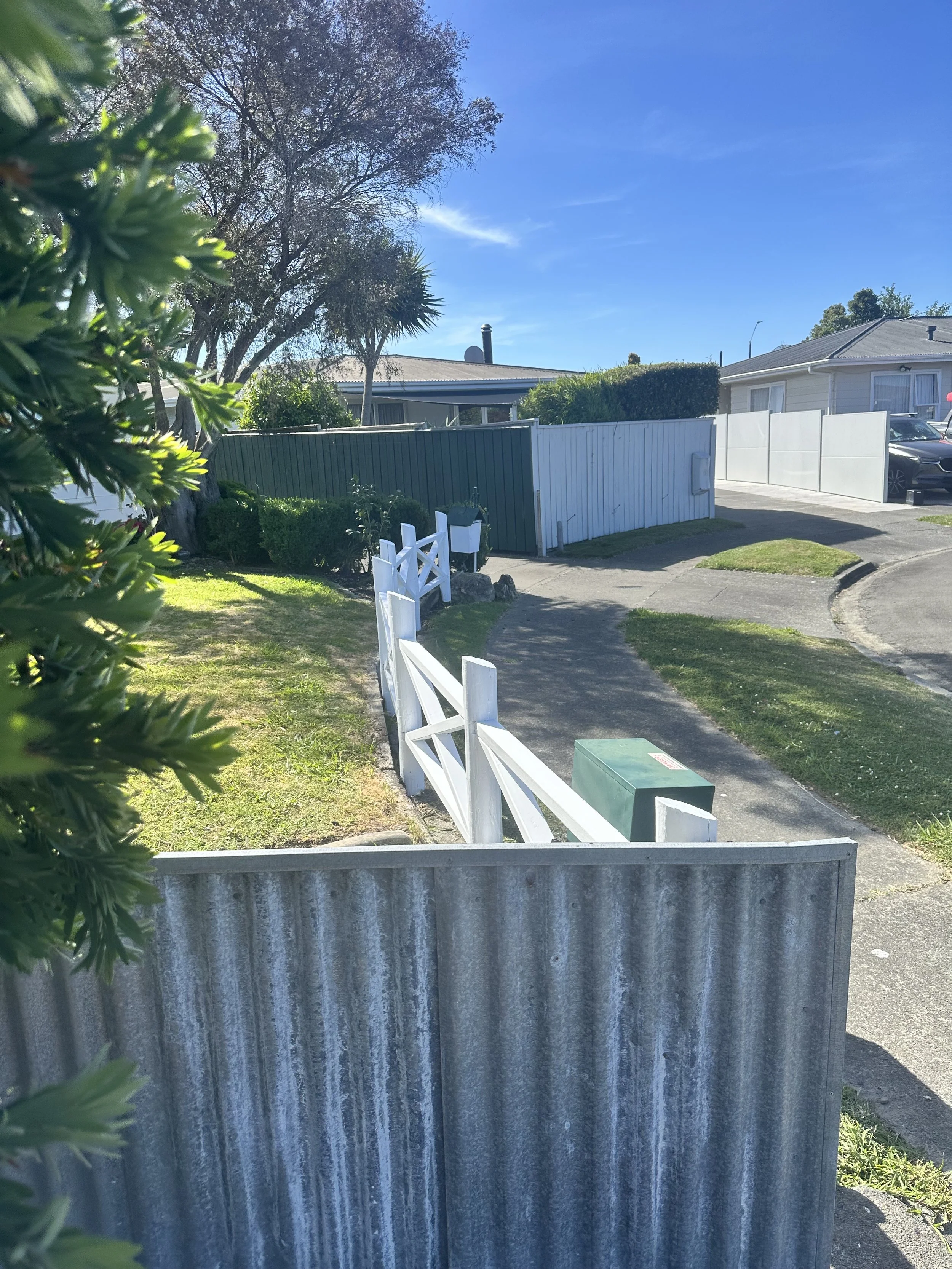 White picket fence freshly sanded and painted white, green hedge, and trees under a clear blue sky.