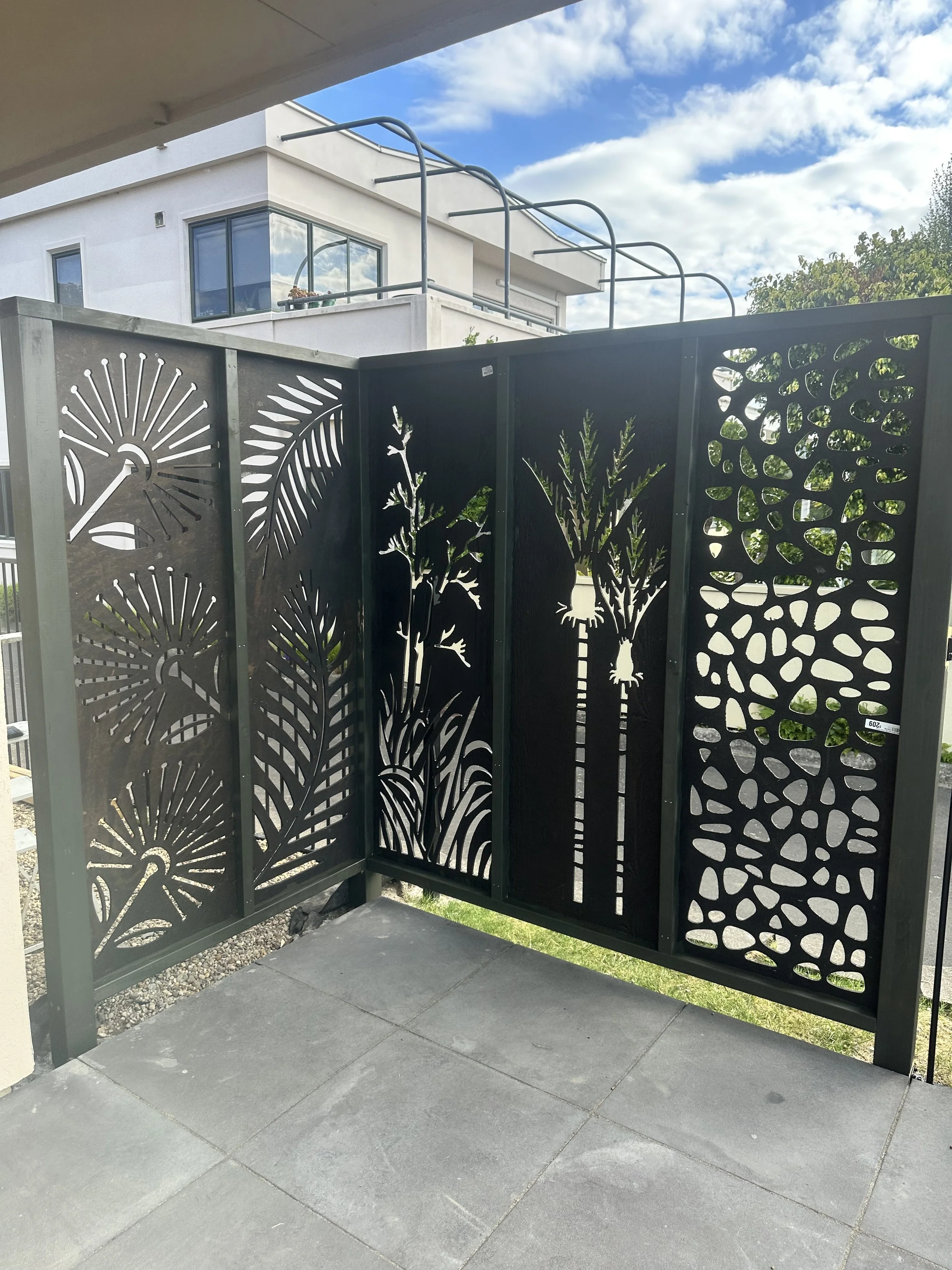 Black decorative metal railing with botanical and abstract patterns on a balcony with a concrete floor, overlooking a modern white building and a cloudy sky.