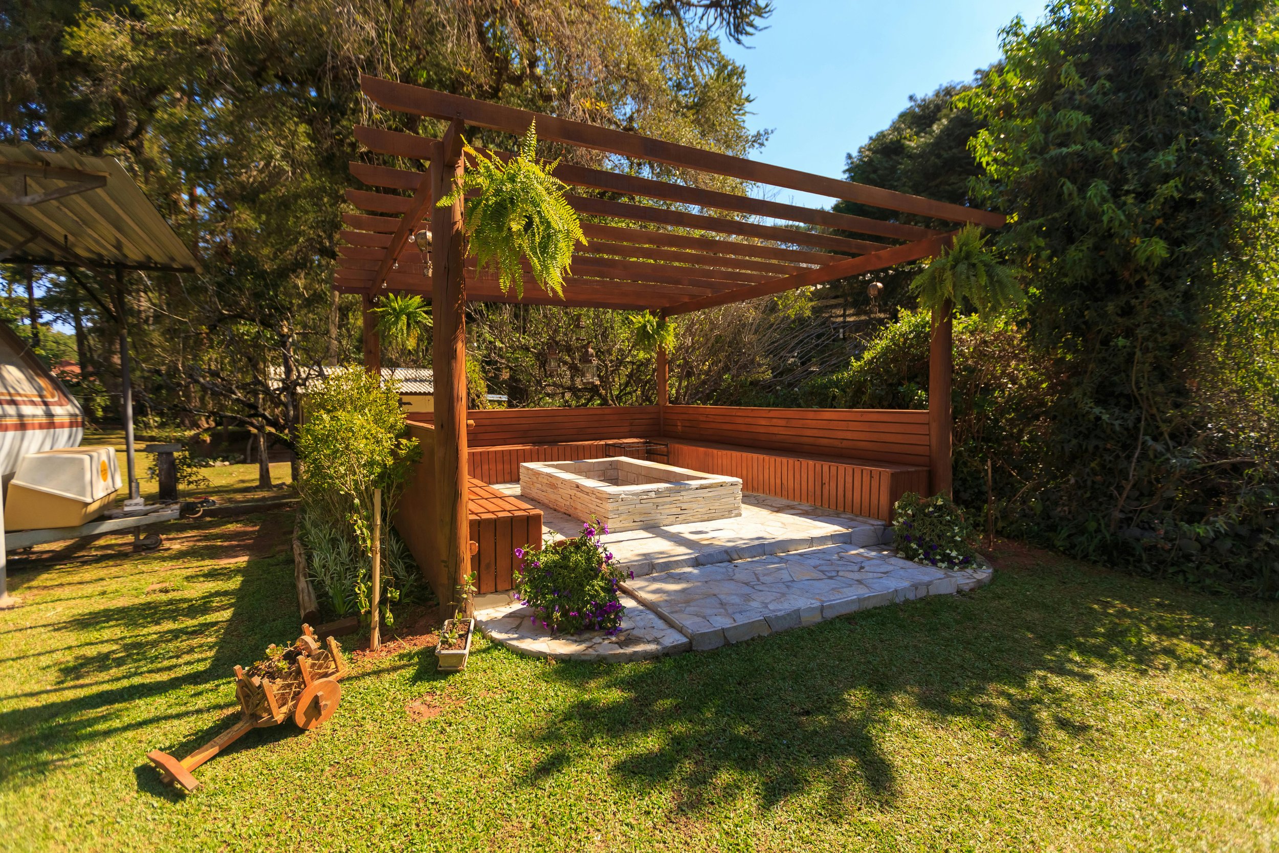 Outdoor backyard patio with a wooden pergola, stone fire pit, and garden plants.