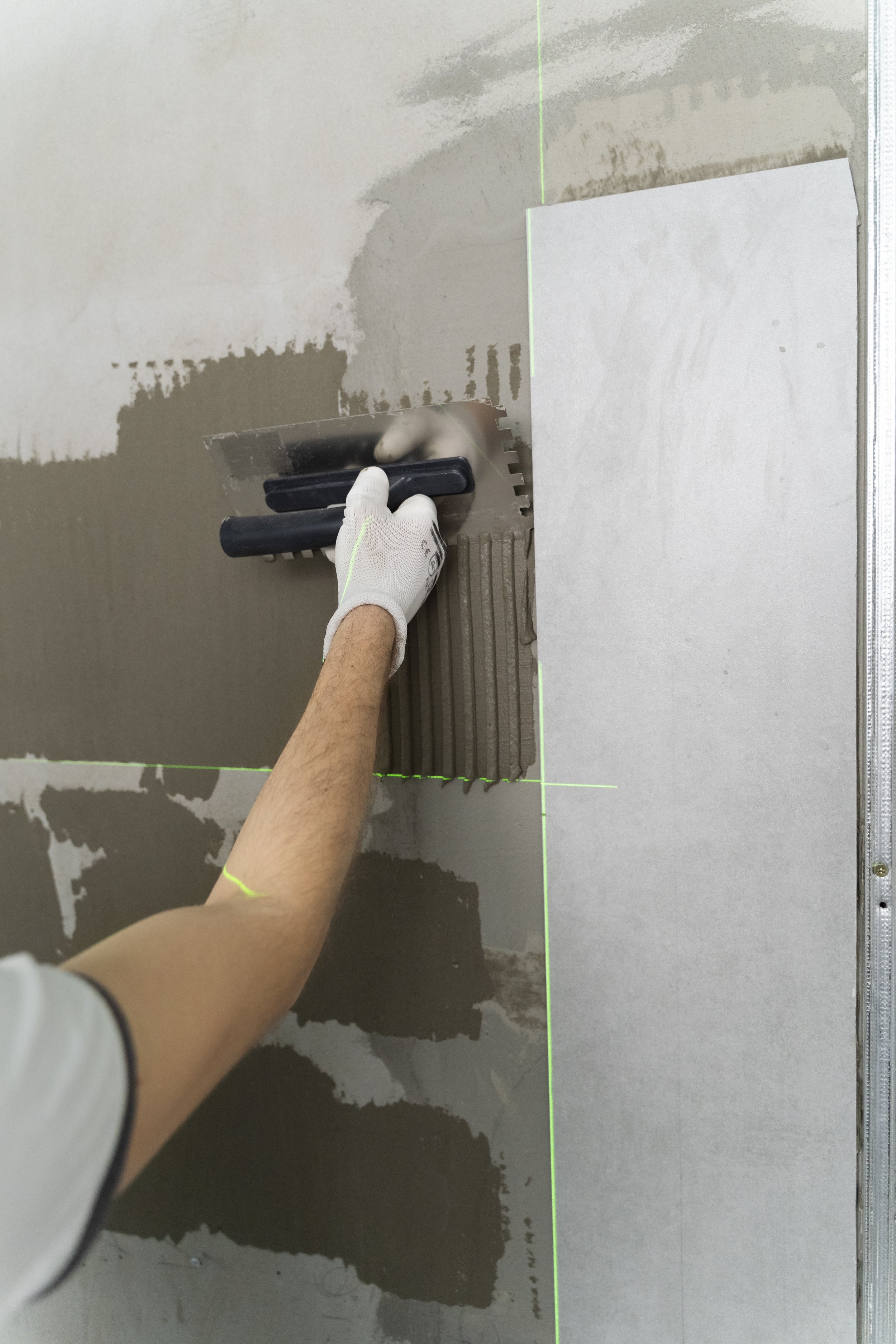 Person wearing white gloves installing drywall with a trowel, smoothing out joint compound in a room under construction, green laser level lines visible.