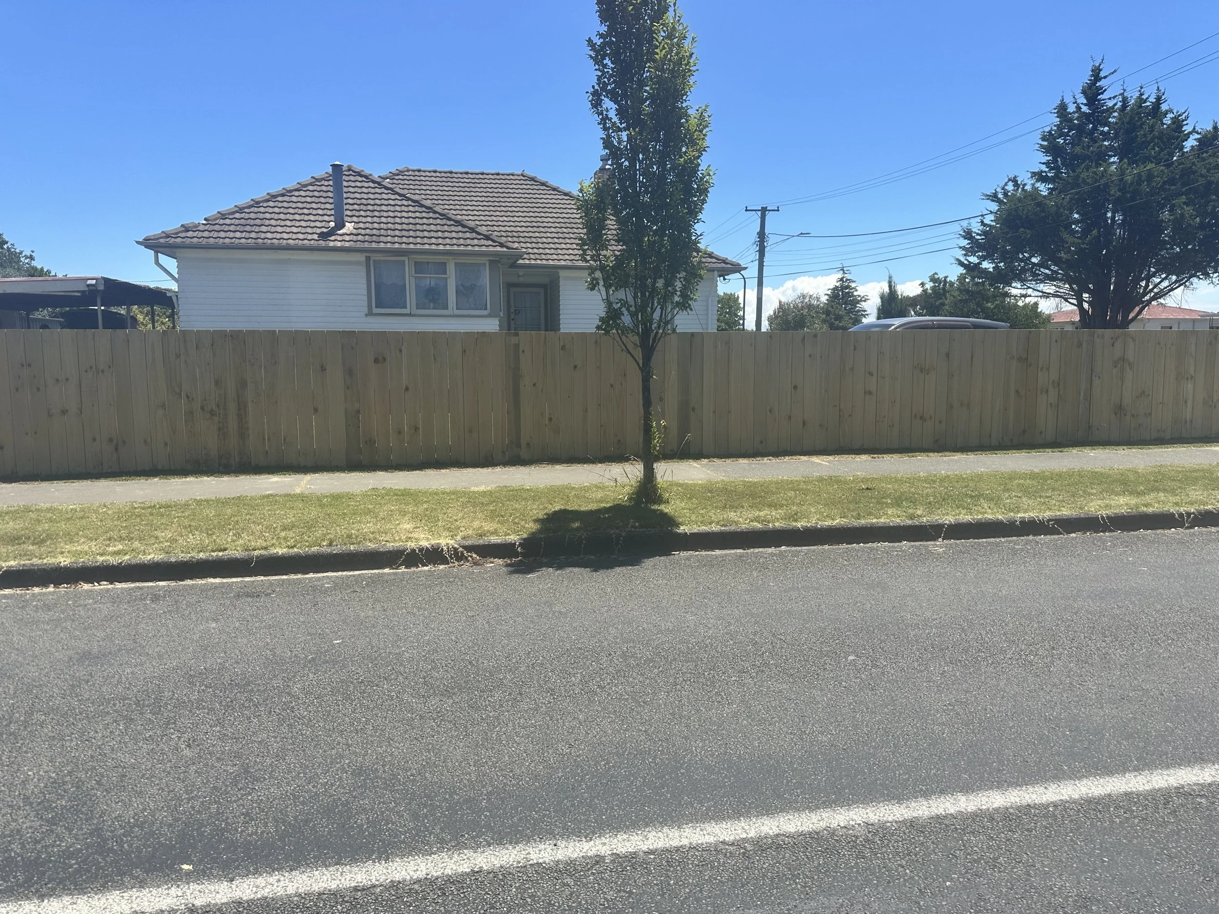 Wooden paling fence, a house with white siding and a brown tiled roof, and a bright blue sky.