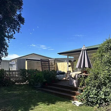 Backyard scene with a deck and outdoor furniture under a large umbrella, surrounded by greenery and fencing, with a clear blue sky above.