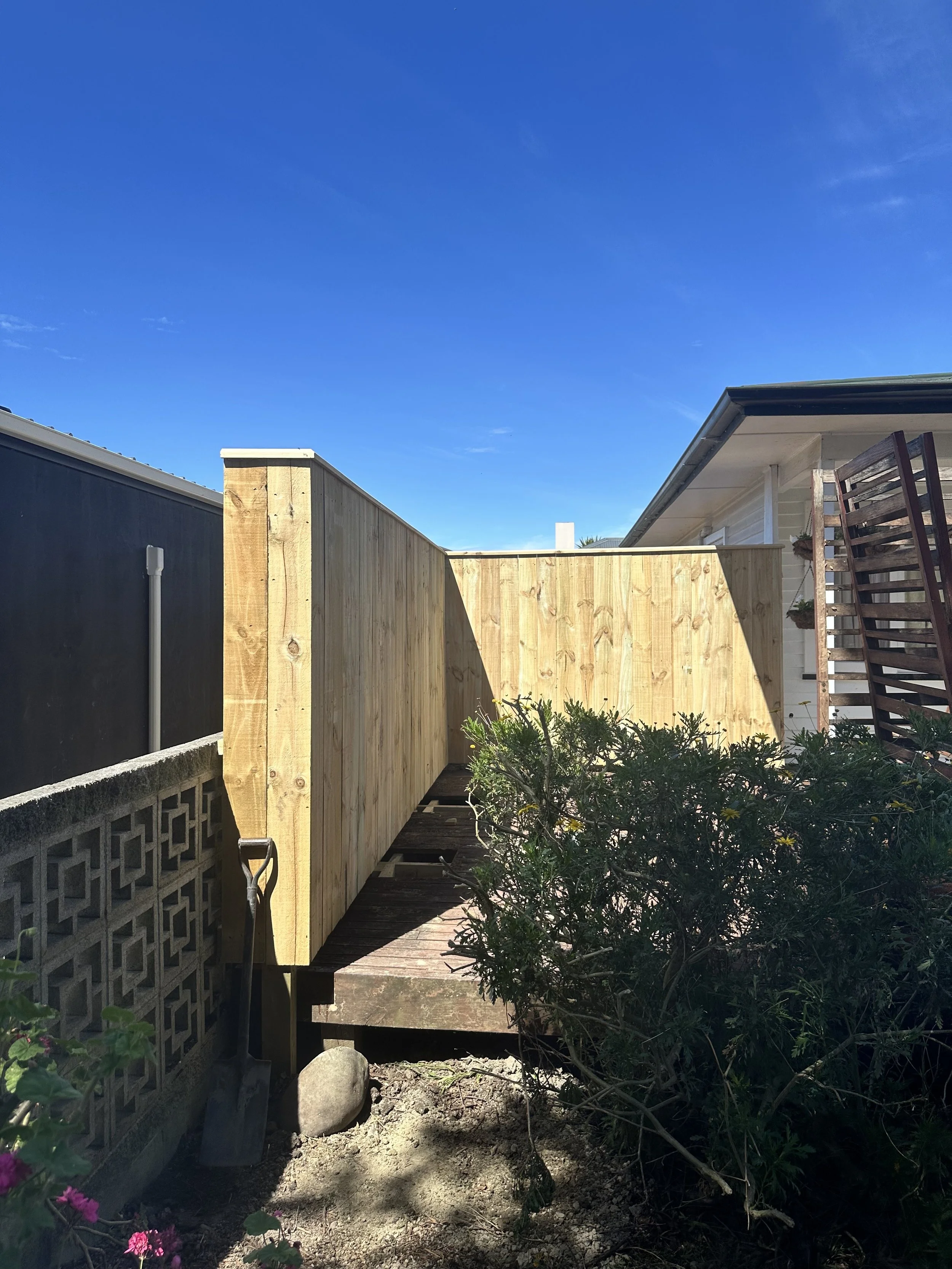 A backyard with a wooden fence under a clear blue sky, with a small garden and a shovel leaning against the wall on the left.