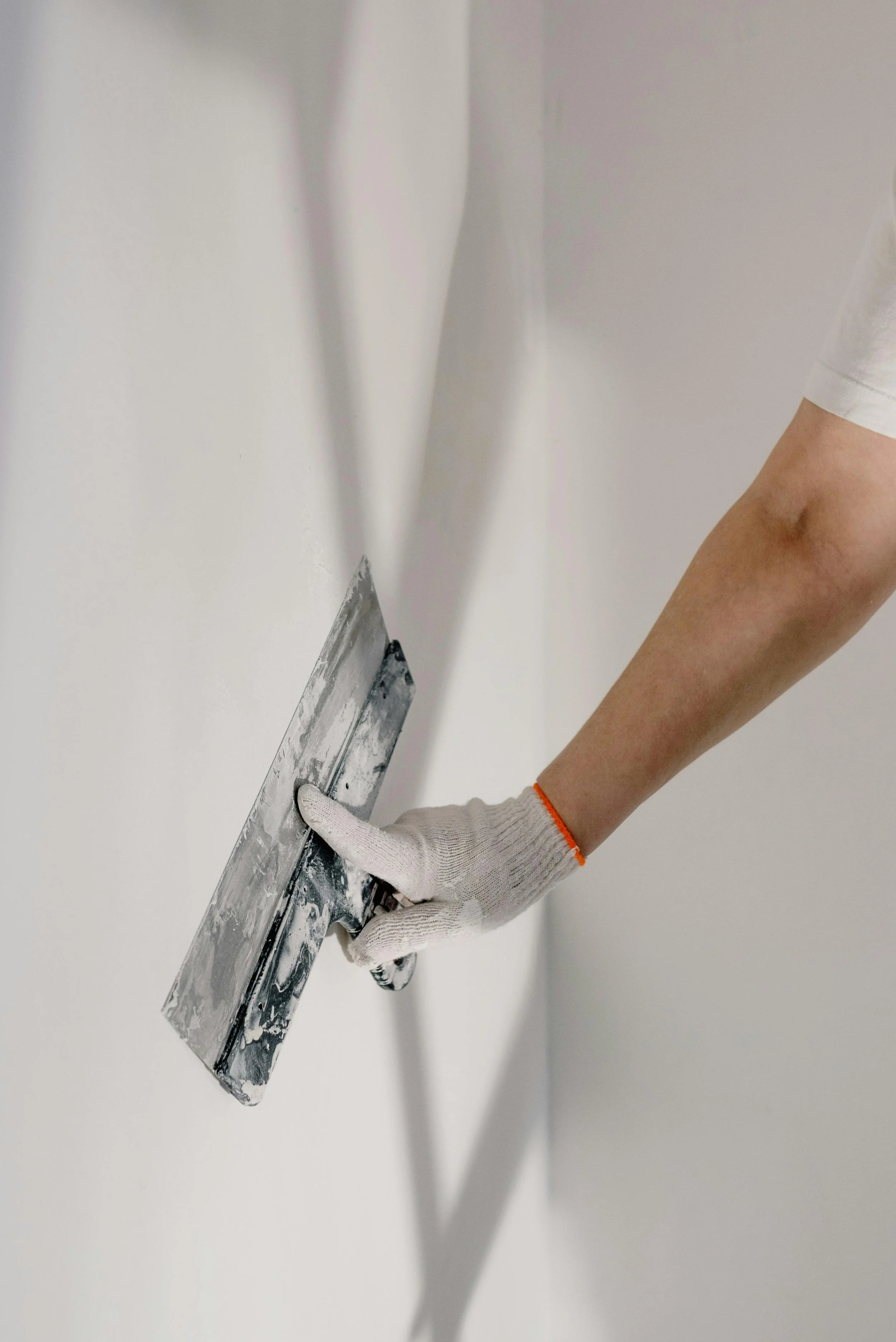 A person with a white glove smoothing fresh white paint on a wall with a large putty knife.