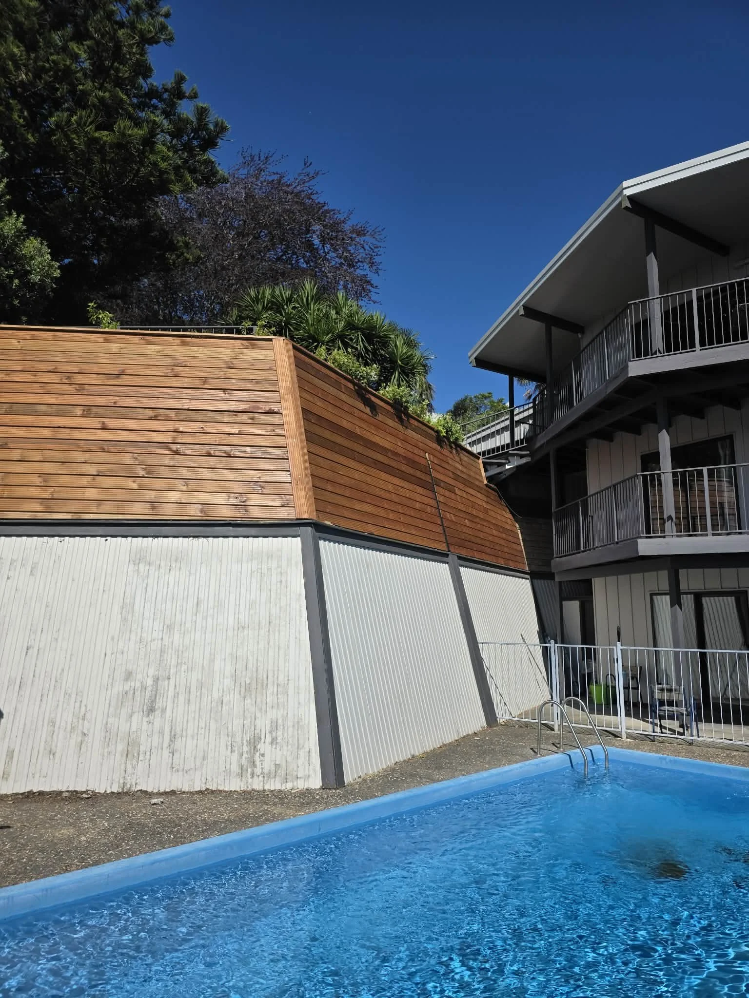 Pool in front of a multi-story residential building with balconies, against a bright blue sky and green trees.