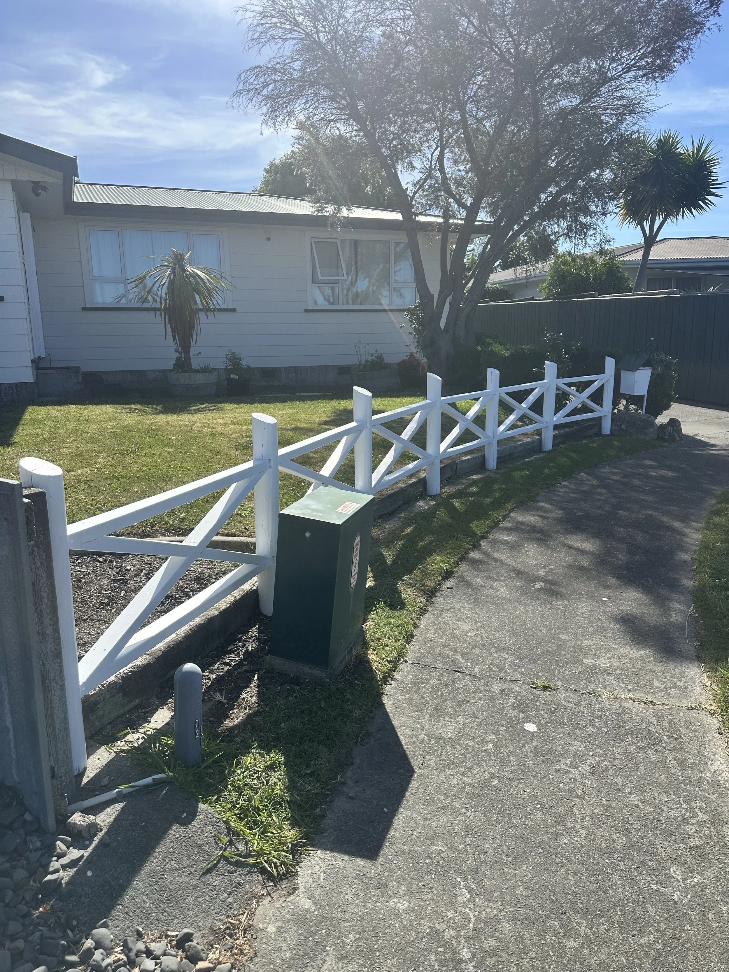 White picket fence freshly sanded and painted in front of a house with a tree and some plants, bathed in sunlight on a clear day.