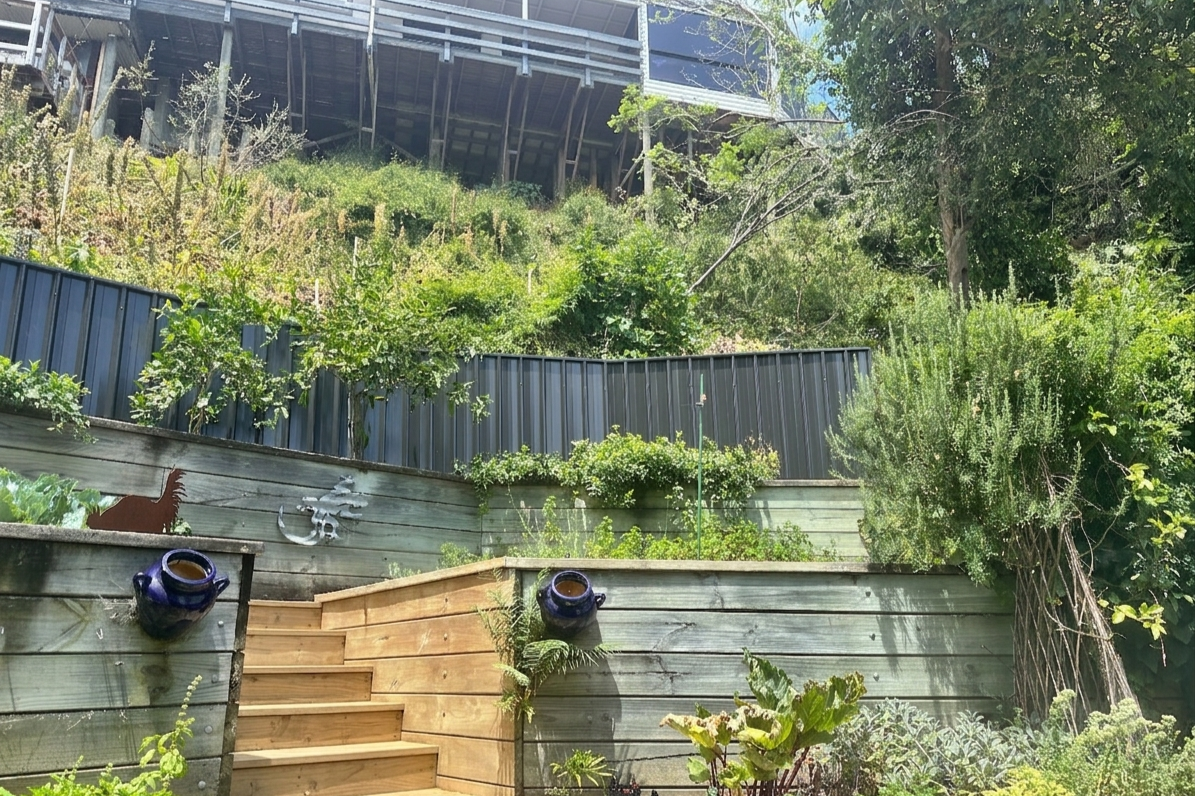 A backyard garden with wooden stairs leading up and a multi-level retaining wall filled with green plants and shrubs, with a house or building partially visible at the top.