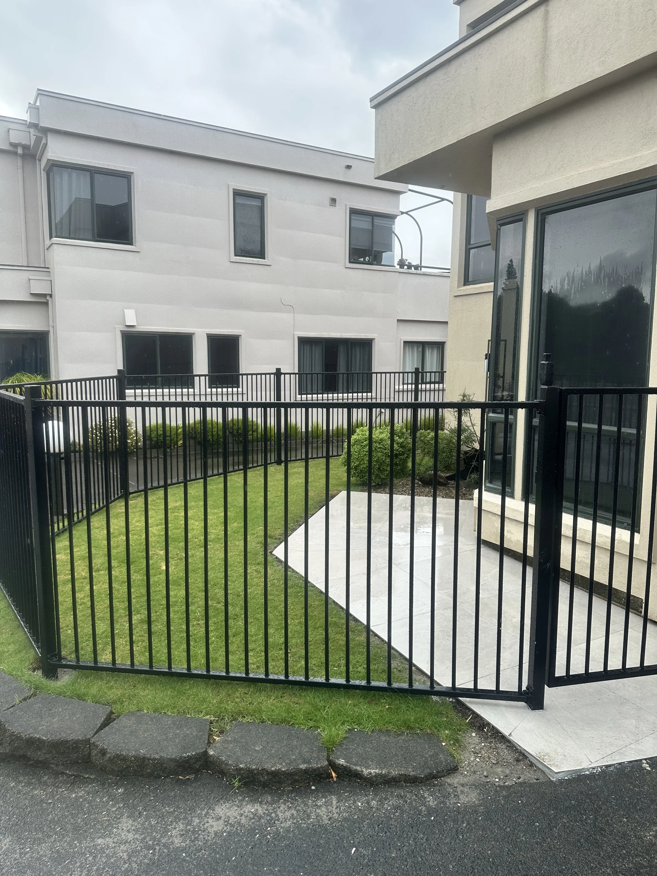 A small fenced yard with a grassy lawn, a concrete patio, and bushes, adjacent to a modern apartment building with large windows.