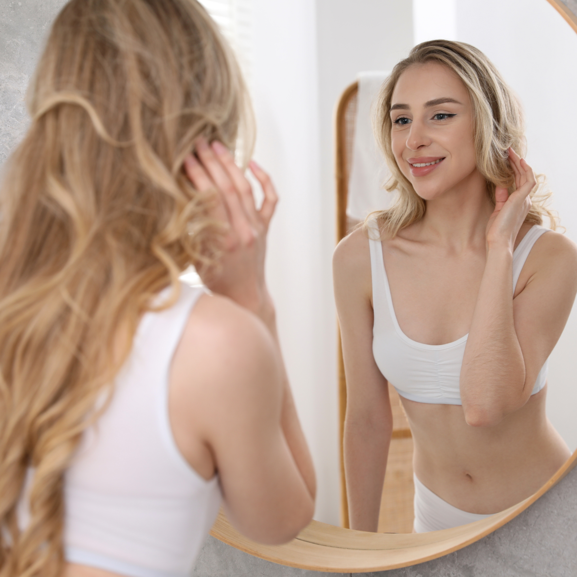A young woman with long wavy blonde hair looking at her reflection in a mirror, smiling softly and touching her hair.