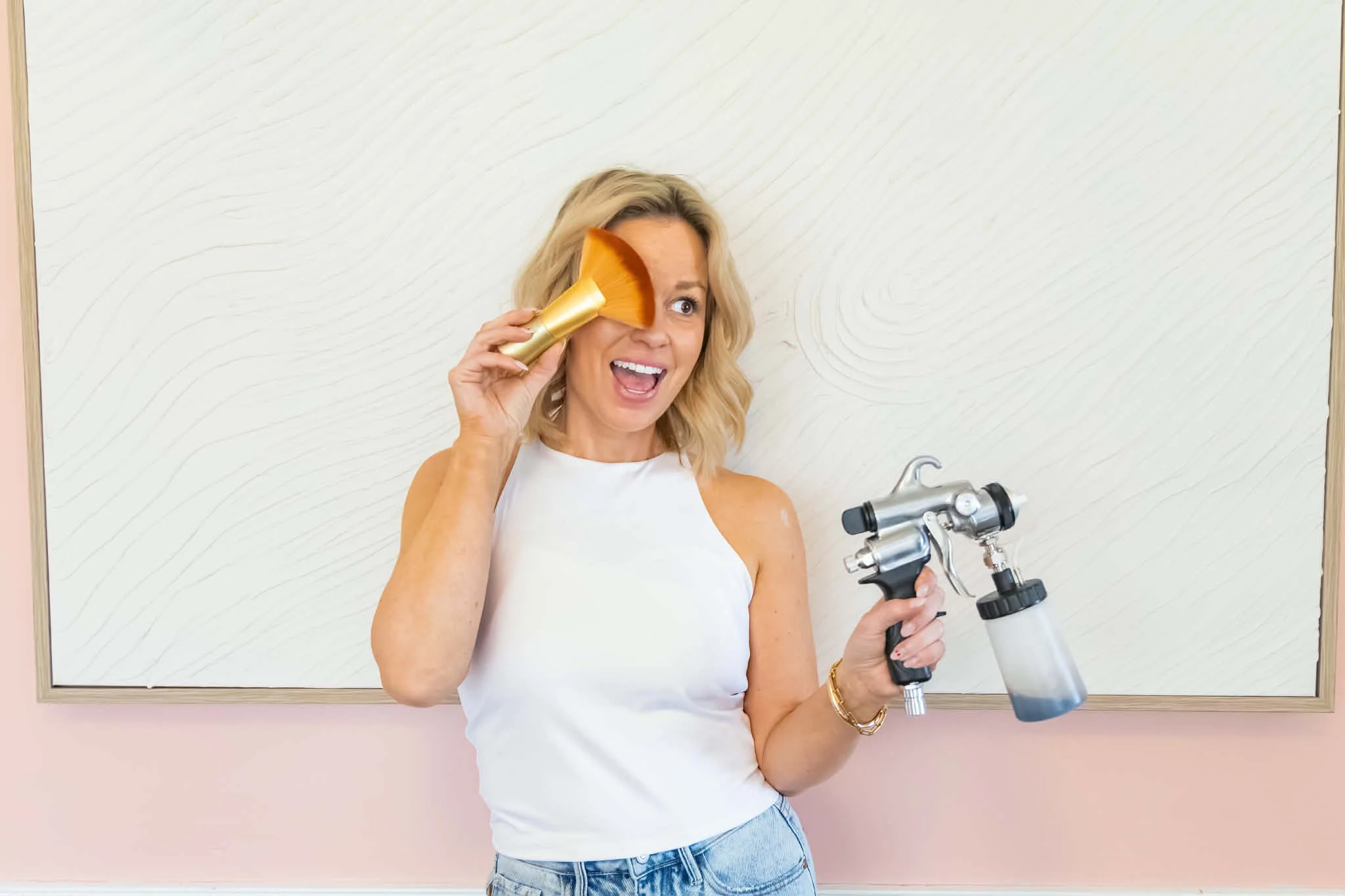 Woman holding a paint spray gun and a paintbrush, smiling and posing in front of a white wall.