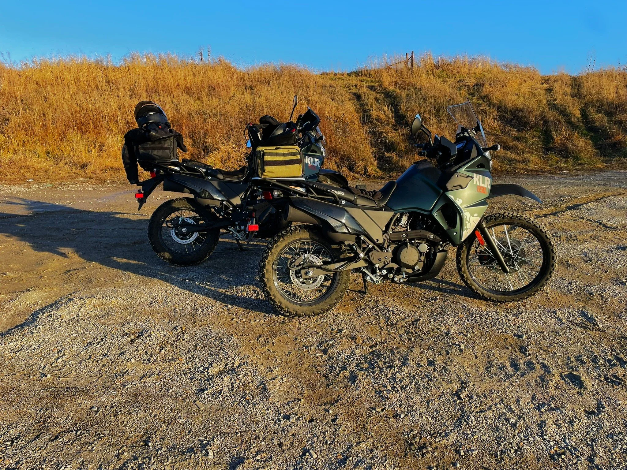 Two adventure motorcycles parked on a dirt surface with dry grass and a clear blue sky in the background.