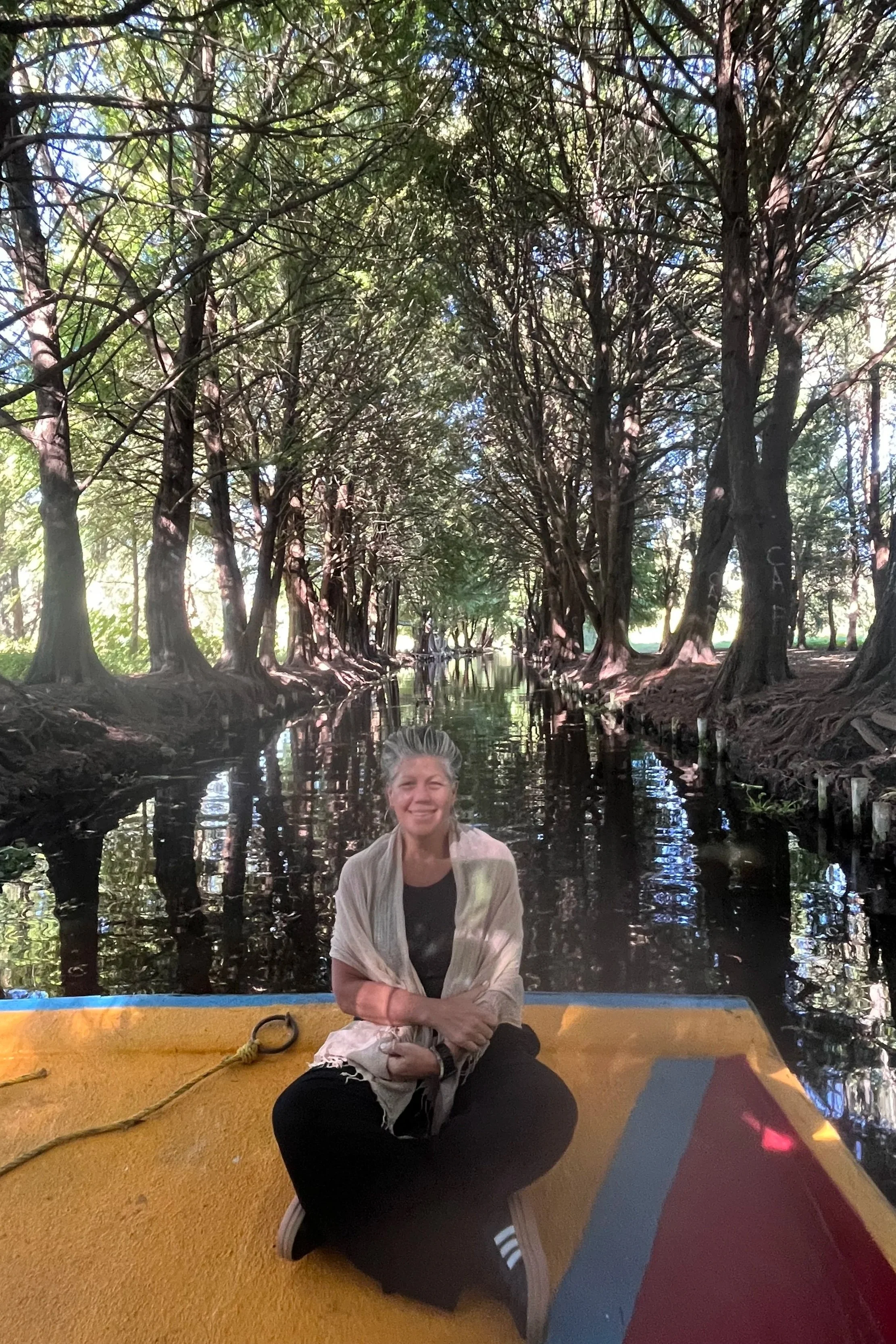 A woman sitting cross-legged on a yellow boat floating on a narrow waterway surrounded by tall trees with dense foliage.