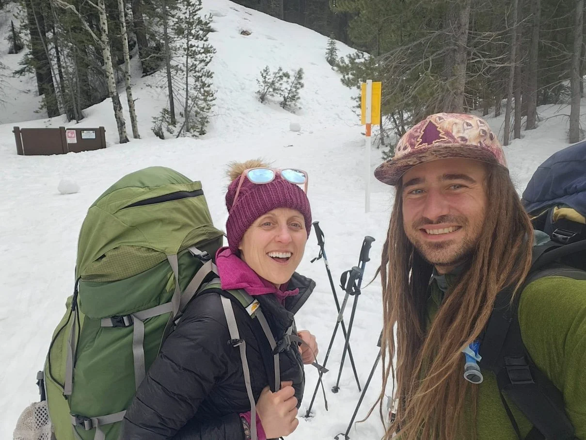Two hikers taking a selfie in a snowy forested area with backpacks, trekking poles, and wearing winter gear.