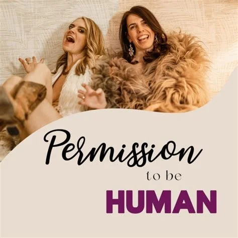 Two women sitting on a bed with a dog, smiling and looking happy, behind a graphic text that reads "Permission to be HUMAN".