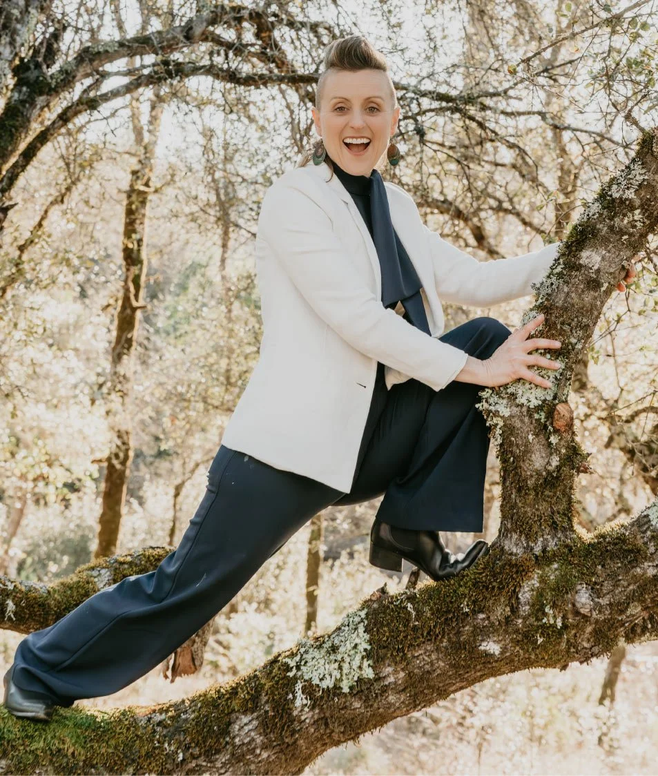 A woman smiling and climbing a tree in an outdoor wooded area during daytime.