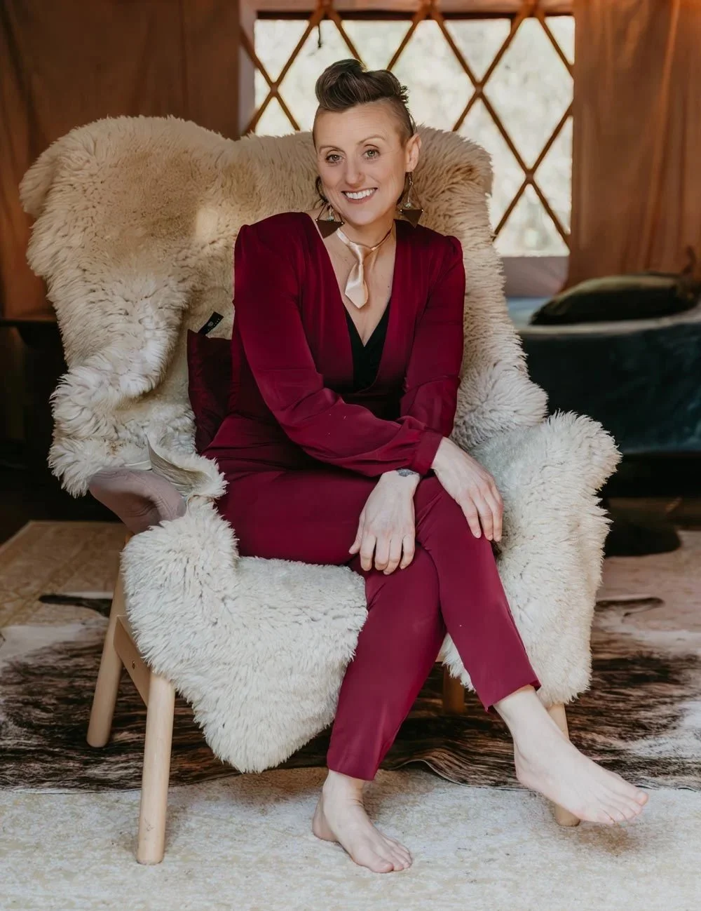 A woman with short, styled hair sitting on a plush, sheepskin-covered chair in a cozy wooden room. She is smiling and wearing a burgundy suit with a beige tiny tie around her neck.