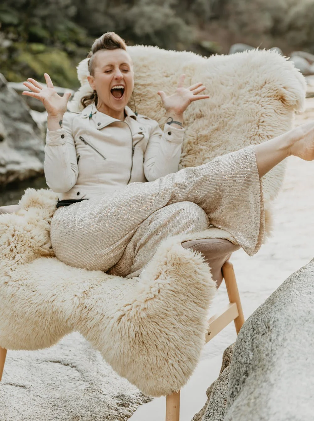 A woman with short hair and a leather jacket sitting on a plush, cream-colored fleece-covered chair outdoors, laughing with her hands in the air, with rocks and blurred greenery in the background.