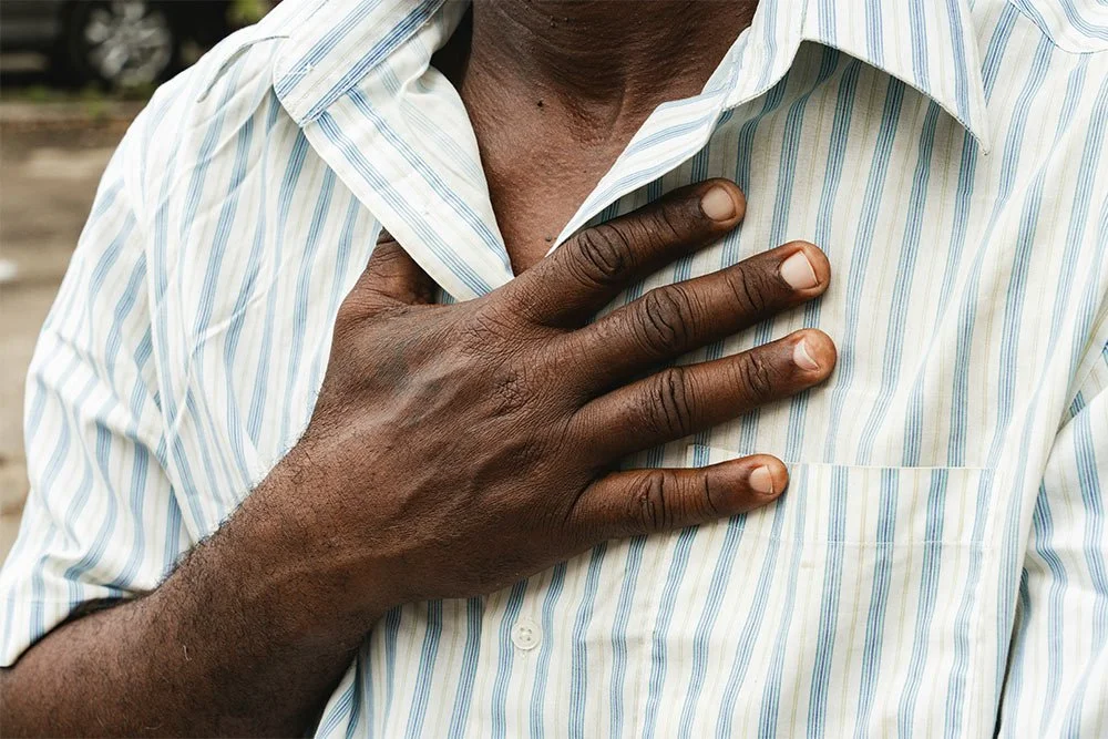 A person with dark skin wearing a light blue and white striped shirt, placing their hand on their chest.