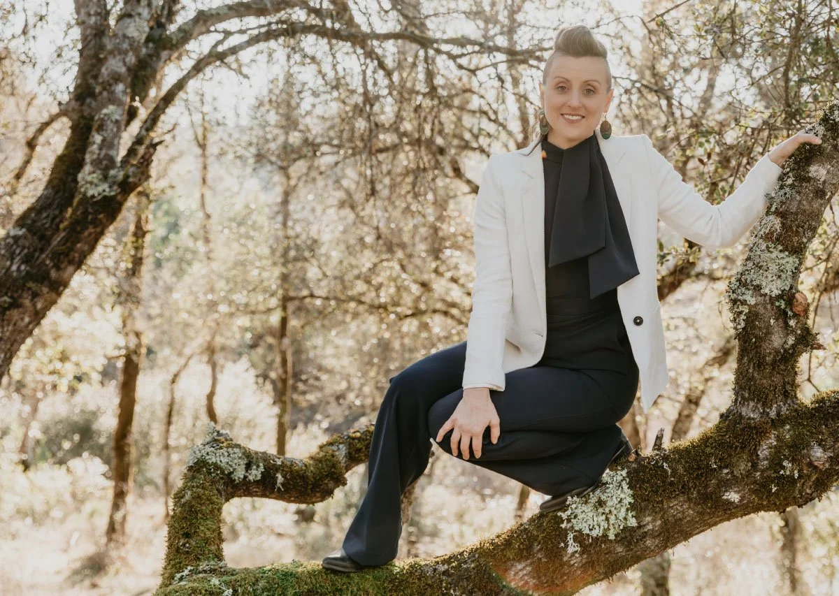 Dr Hazel-Grace smiling, crouched in a tree in a white blazer and navy pants