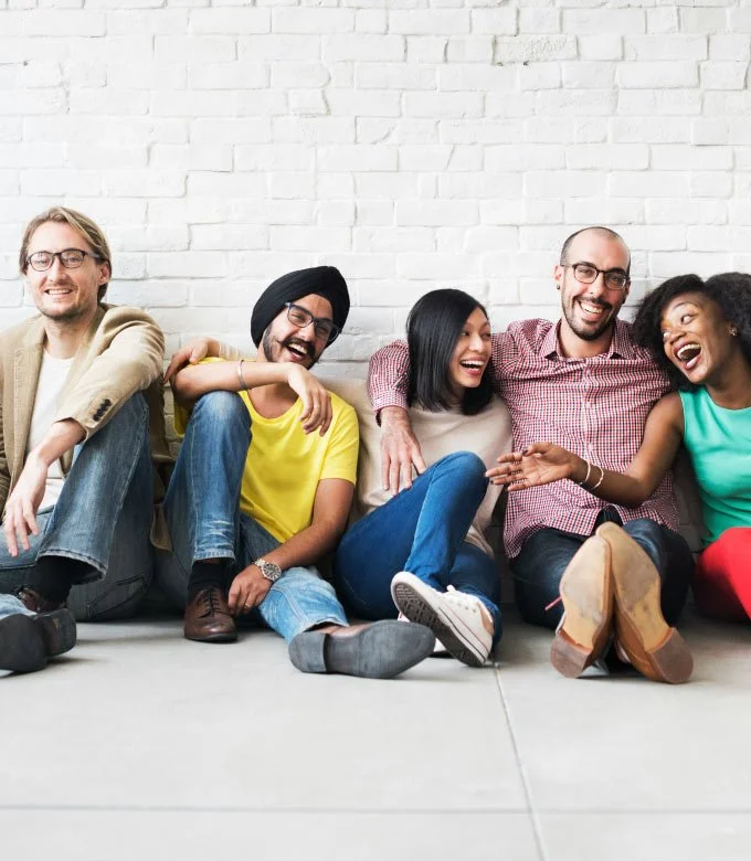 Group of men and women of different ethnicities wearing colorful clothes sitting on ground against white brick wall