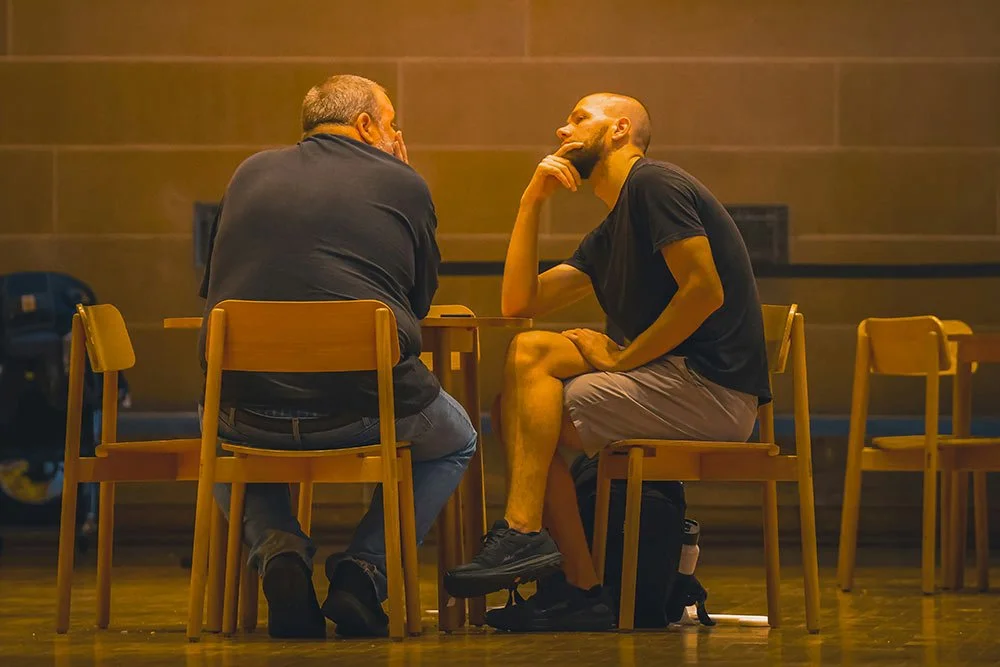Two men sitting across from each other at a table, one older and one younger, engaged in a conversation, in a warm-toned indoor setting.