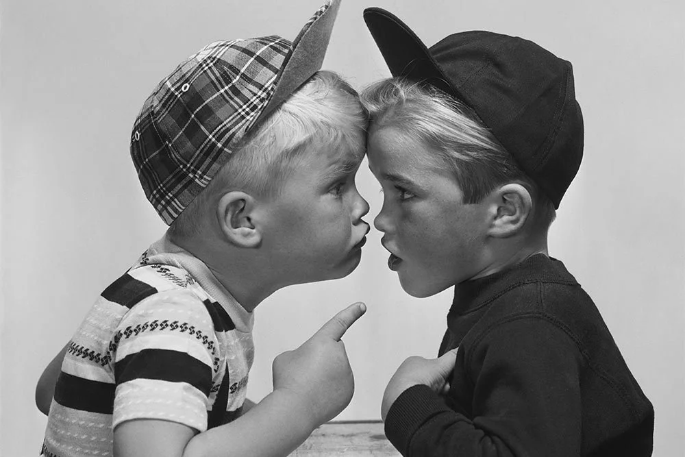 Two young boys face each other closely, with their foreheads almost touching, in a playful confrontation. The boy on the left wears a baseball cap with a plaid pattern, and the boy on the right wears a plain cap. One boy points his finger at the other, and both boys have serious expressions in a black-and-white photo.