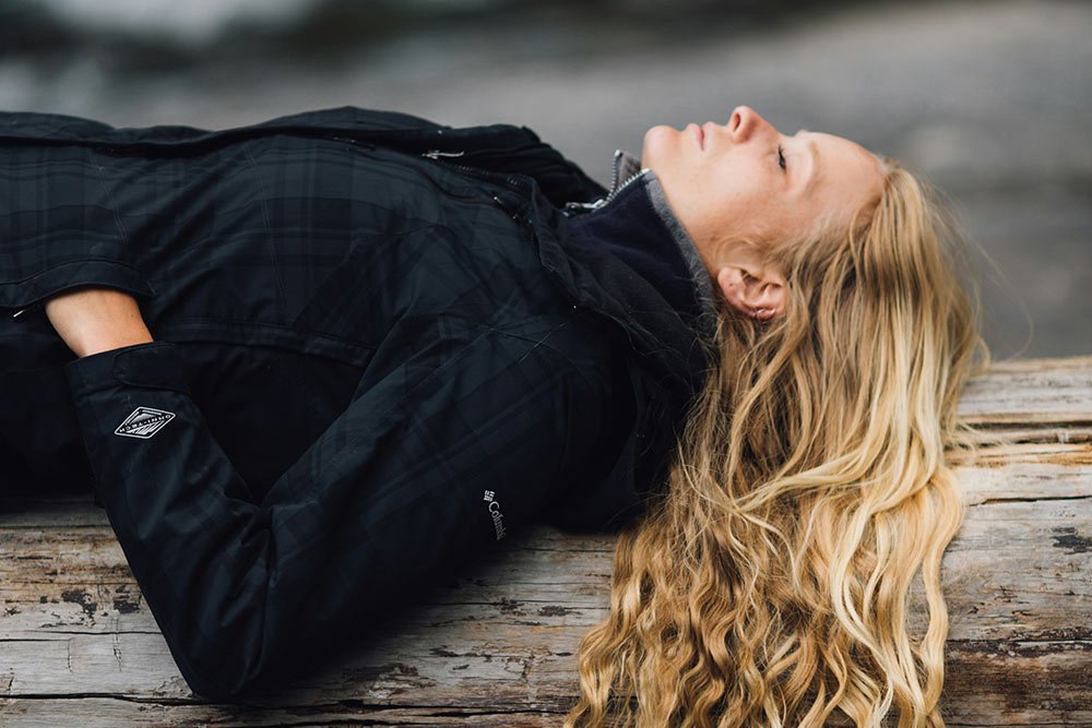 Woman lying on her back on a wooden dock with her eyes closed, wearing a black jacket and a dark shirt, with long, wavy blonde hair.