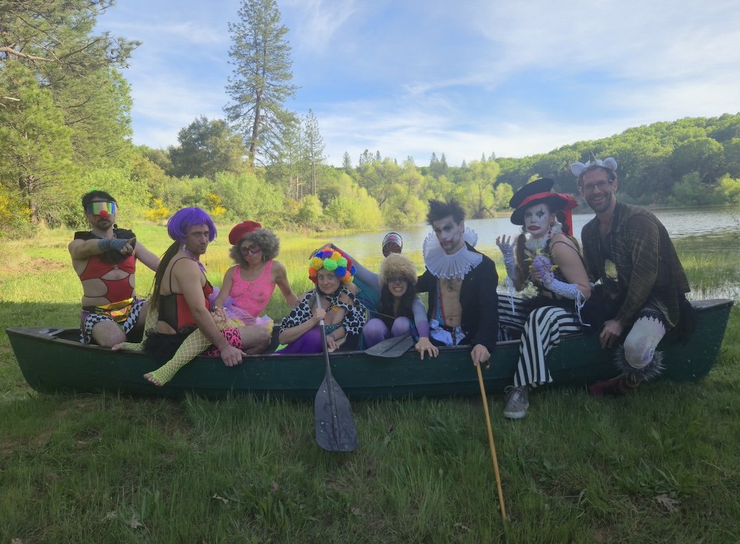 People dressed as clowns, a mime, and a frog in a boat on a lake with trees and blue sky in the background.